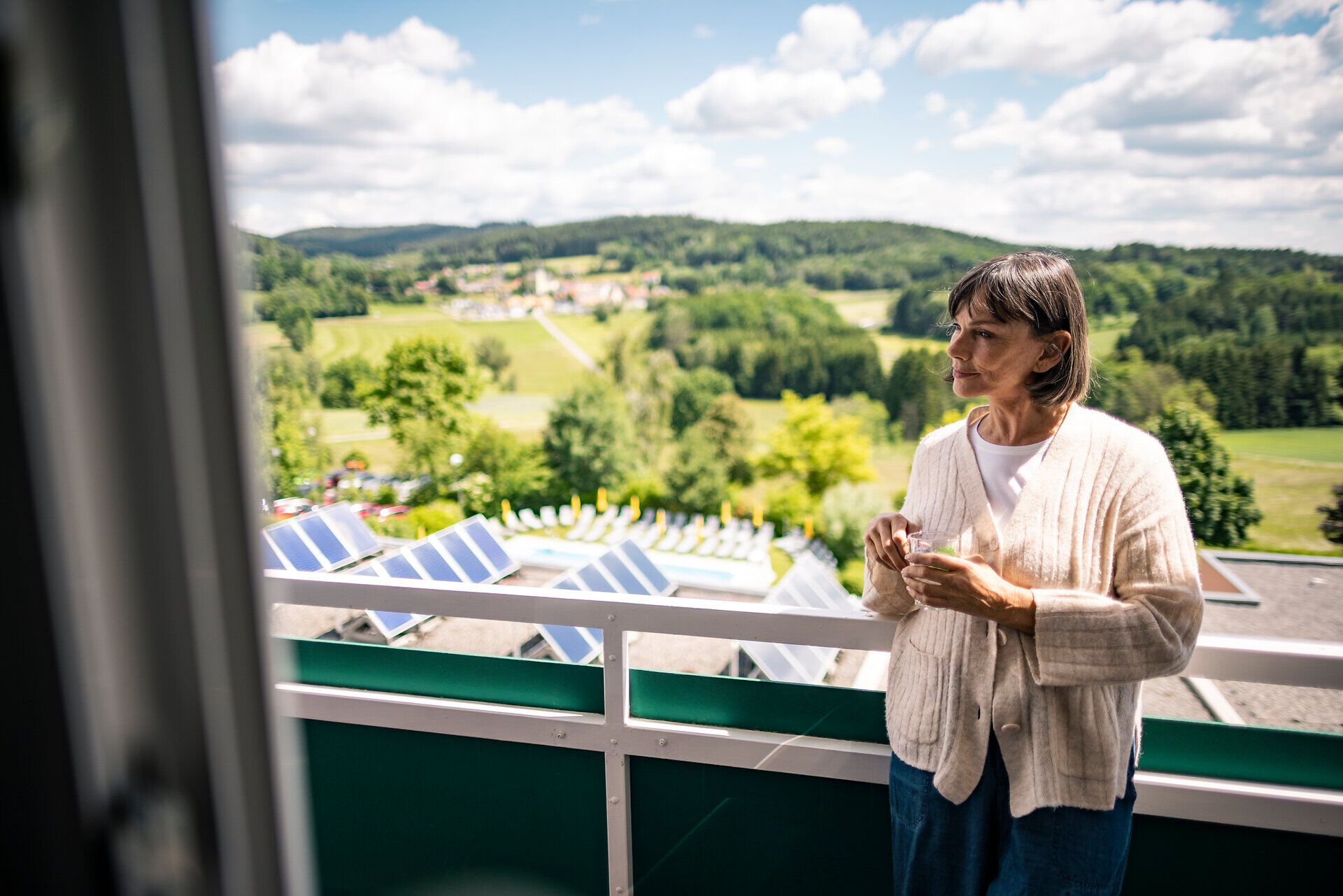 Osoba s šálkem na balkóně zdravotního a rehabilitačního centra Moorheilbad Harbach ve Waldviertelu s výhledem na zelenou kopcovitou krajinu.