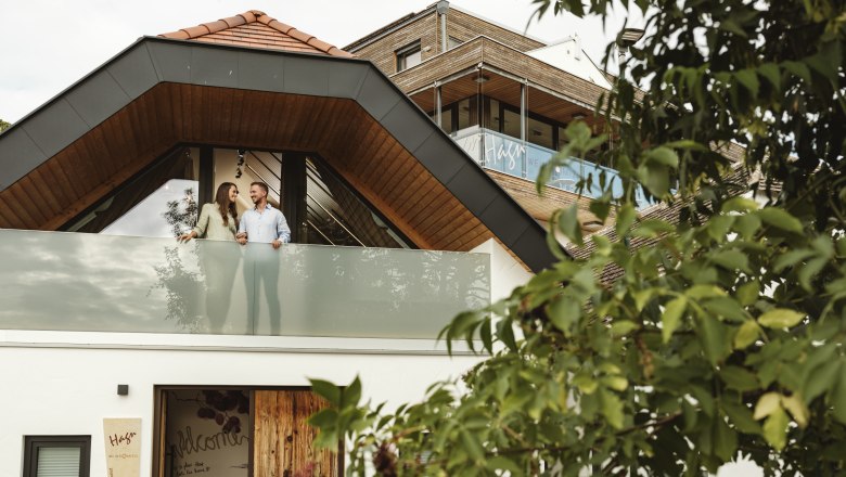 Modern&iacute; budova s p&aacute;rem na balkoně, obklopen&aacute; stromy.