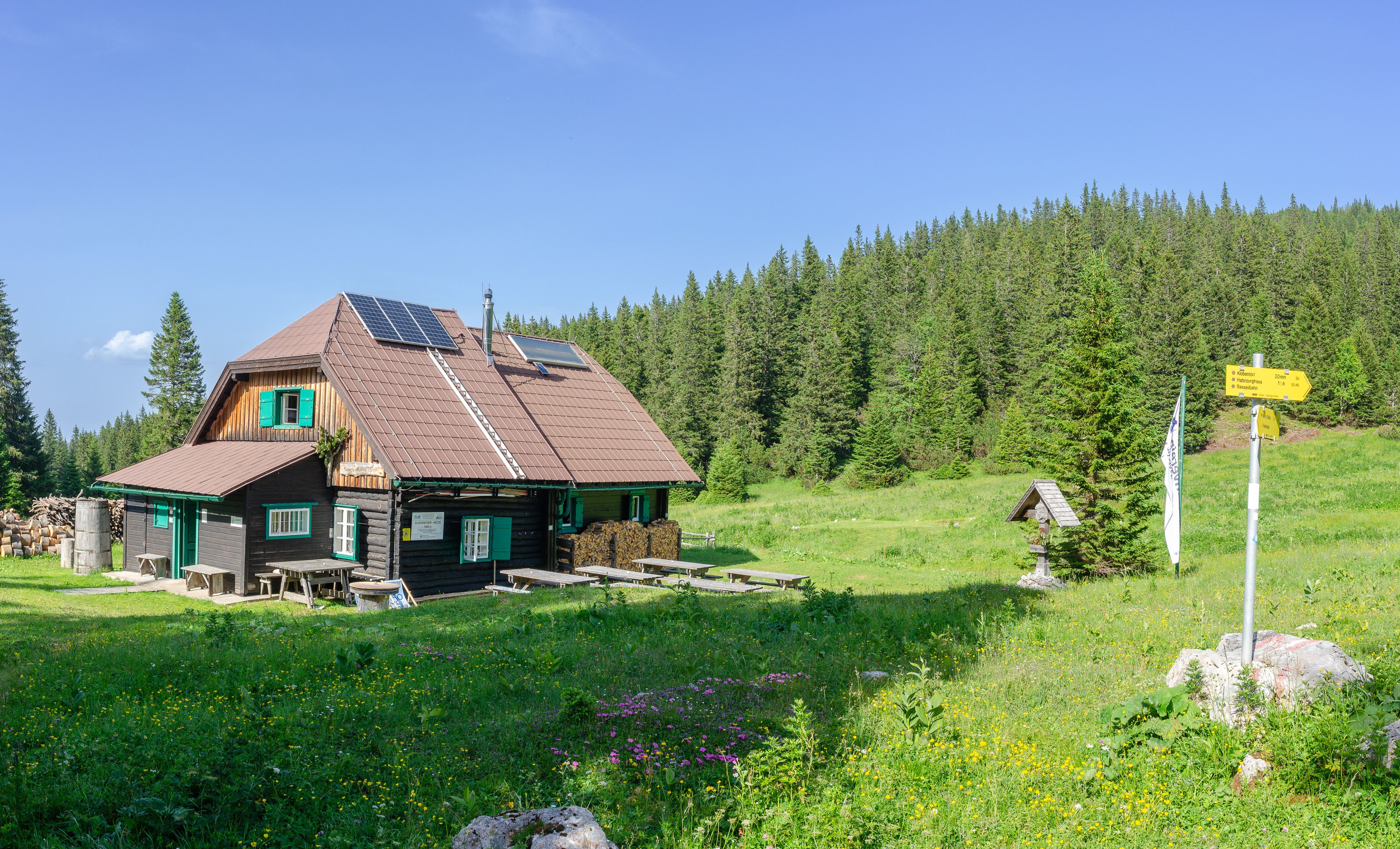 Horská chata se solárními panely na střeše, obklopená zelenou loukou a lesem.