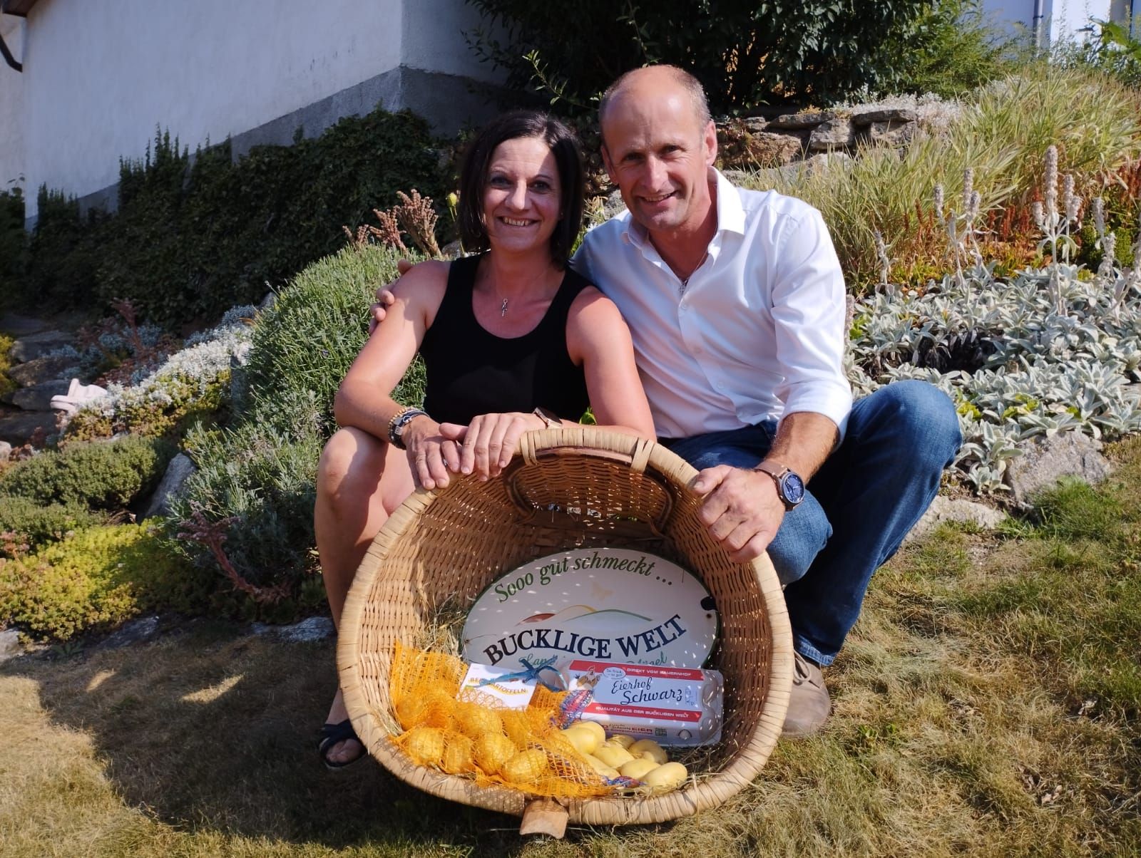 Žena a muž sedí s úsměvem na zahradě a drží košík s bramborami a vejci.