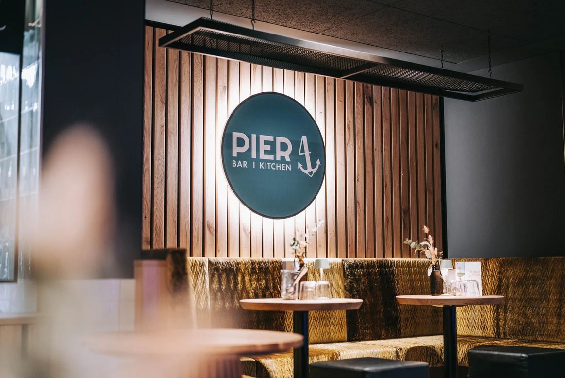 Pohled do interiéru restaurace Pier 4 s dřevěnou stěnou a logem.
