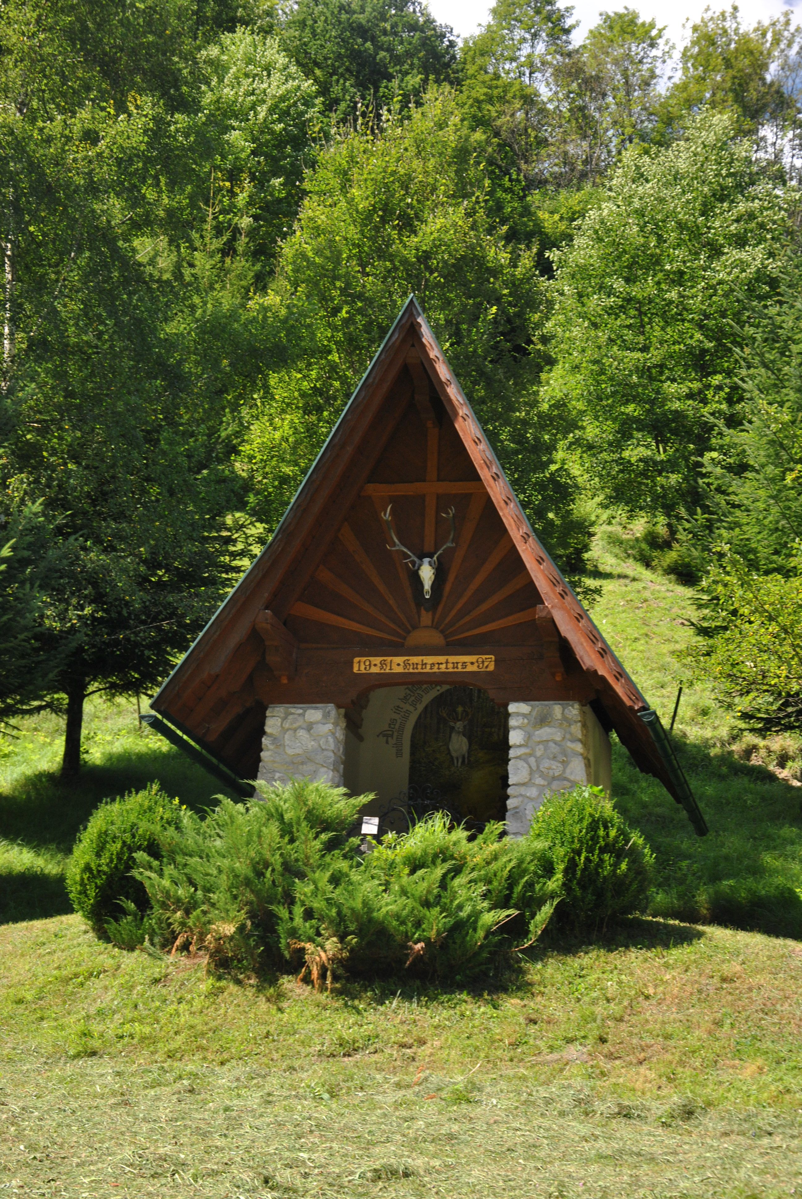 Malá kaple se špičatou střechou a jeleními parohy, obklopená stromy a keři.