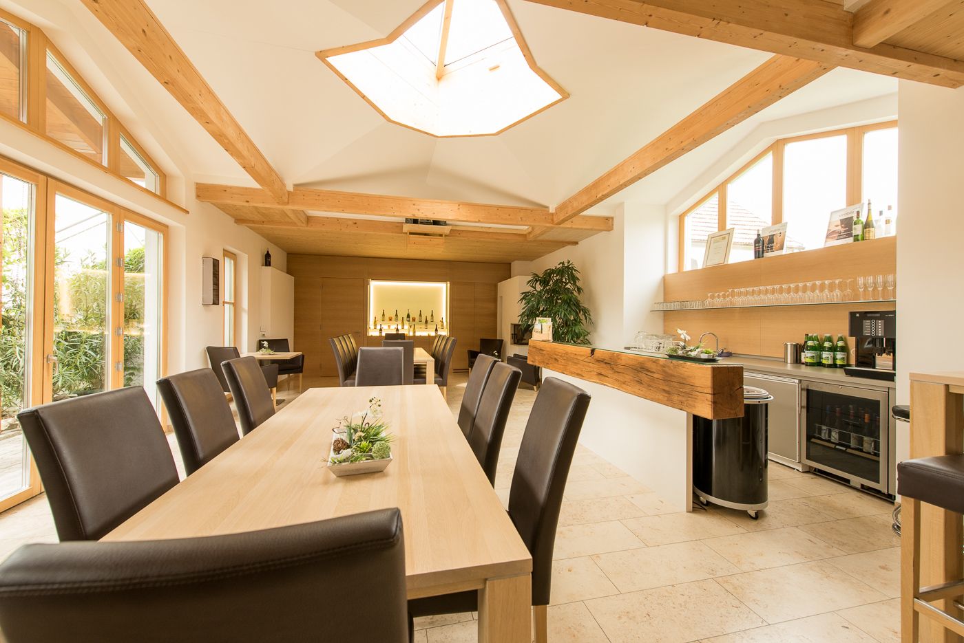 Pohled do interiéru moderní degustační místnosti s dřevěnými stoly a židlemi, velkými okny a barem s lahvemi vína.