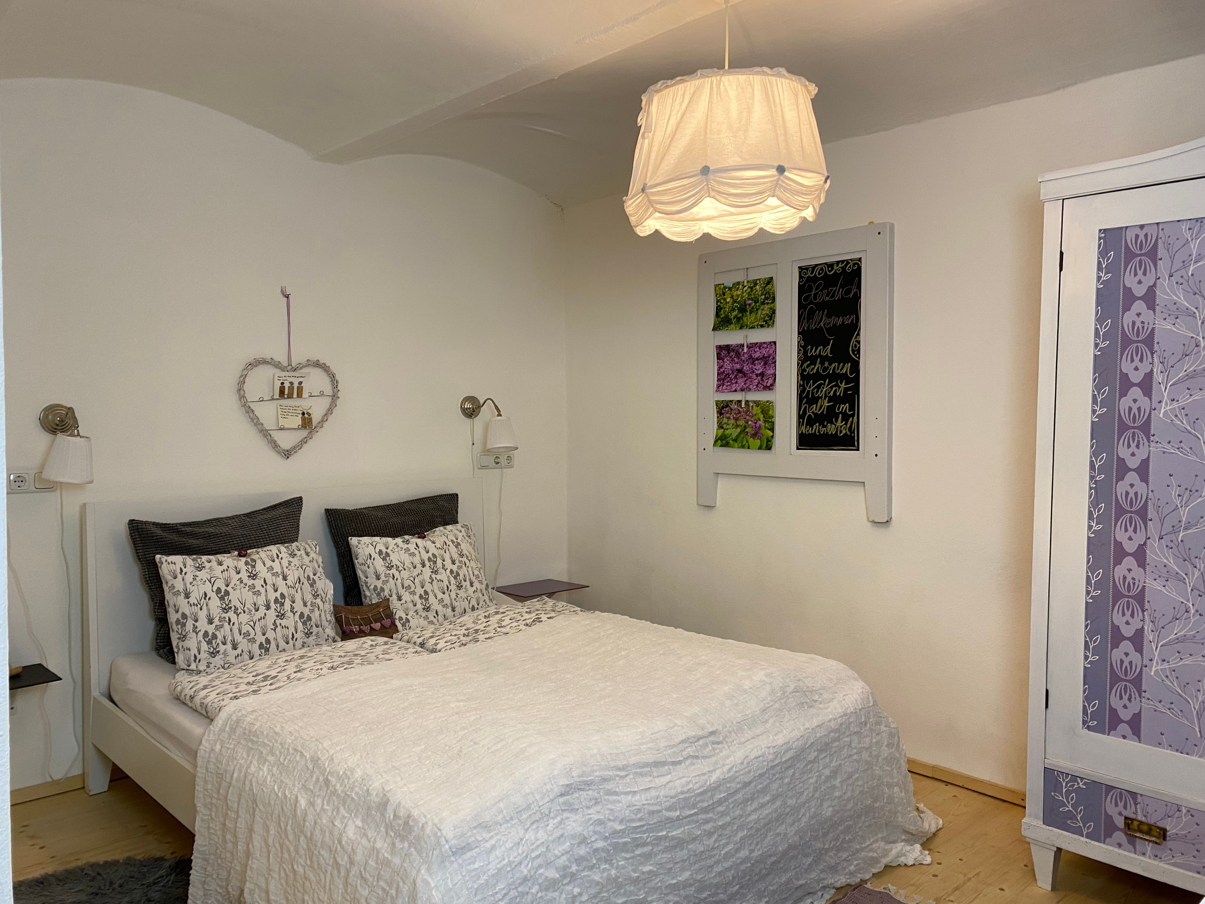 Útulný dvoulůžkový pokoj s bílou postelí, ozdobnou policí se srdcem a fialovou šatní skříní.