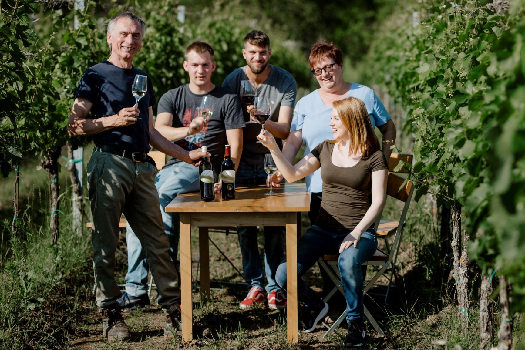 U stolu na vinici sedí a stojí skupina pěti lidí, v rukou drží sklenky s vínem a usmívají se.
