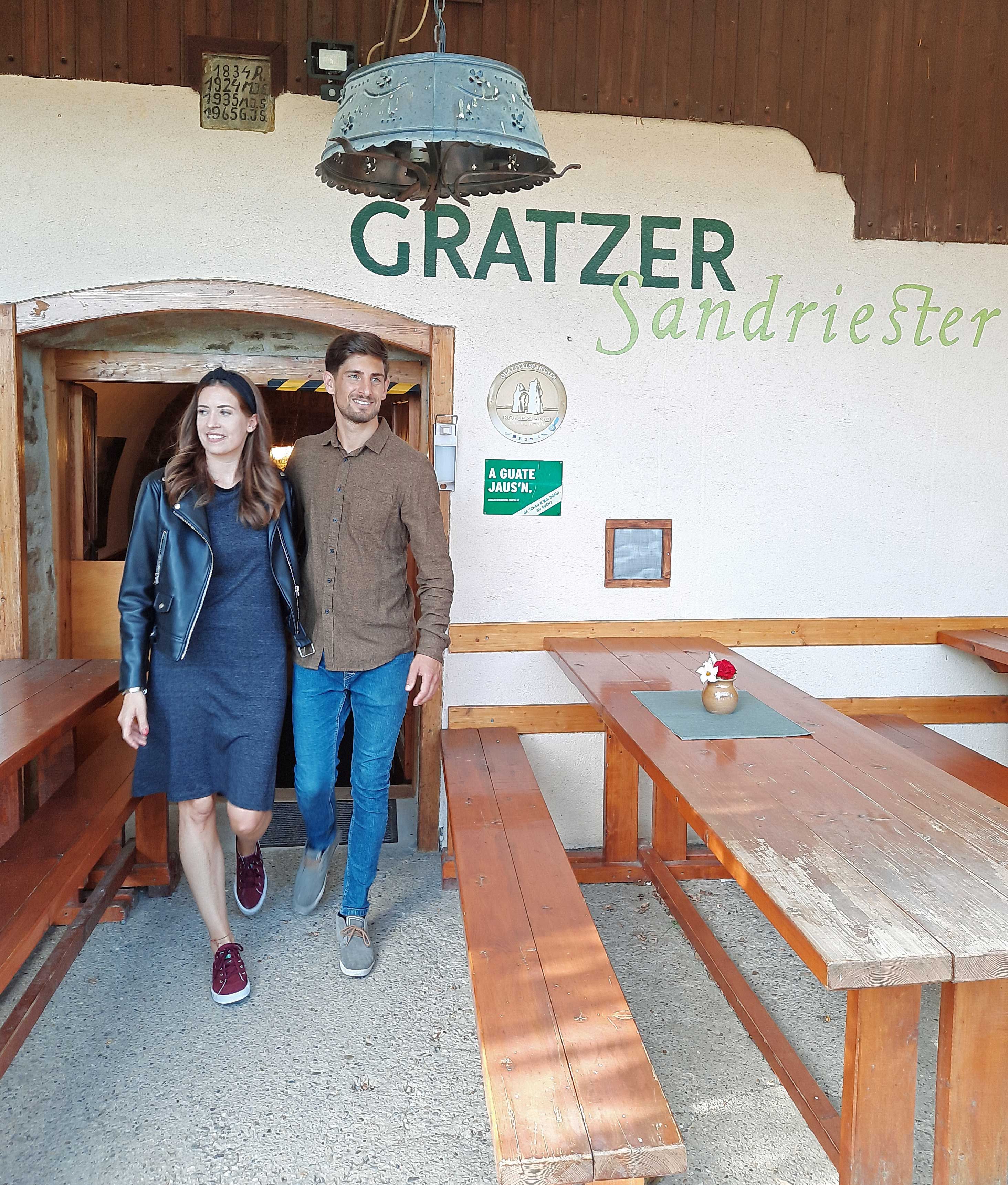 Muž a žena odcházejí z vinného šenku Gratzer-Sandriester. Usmívají se a procházejí kolem dřevěných lavic.