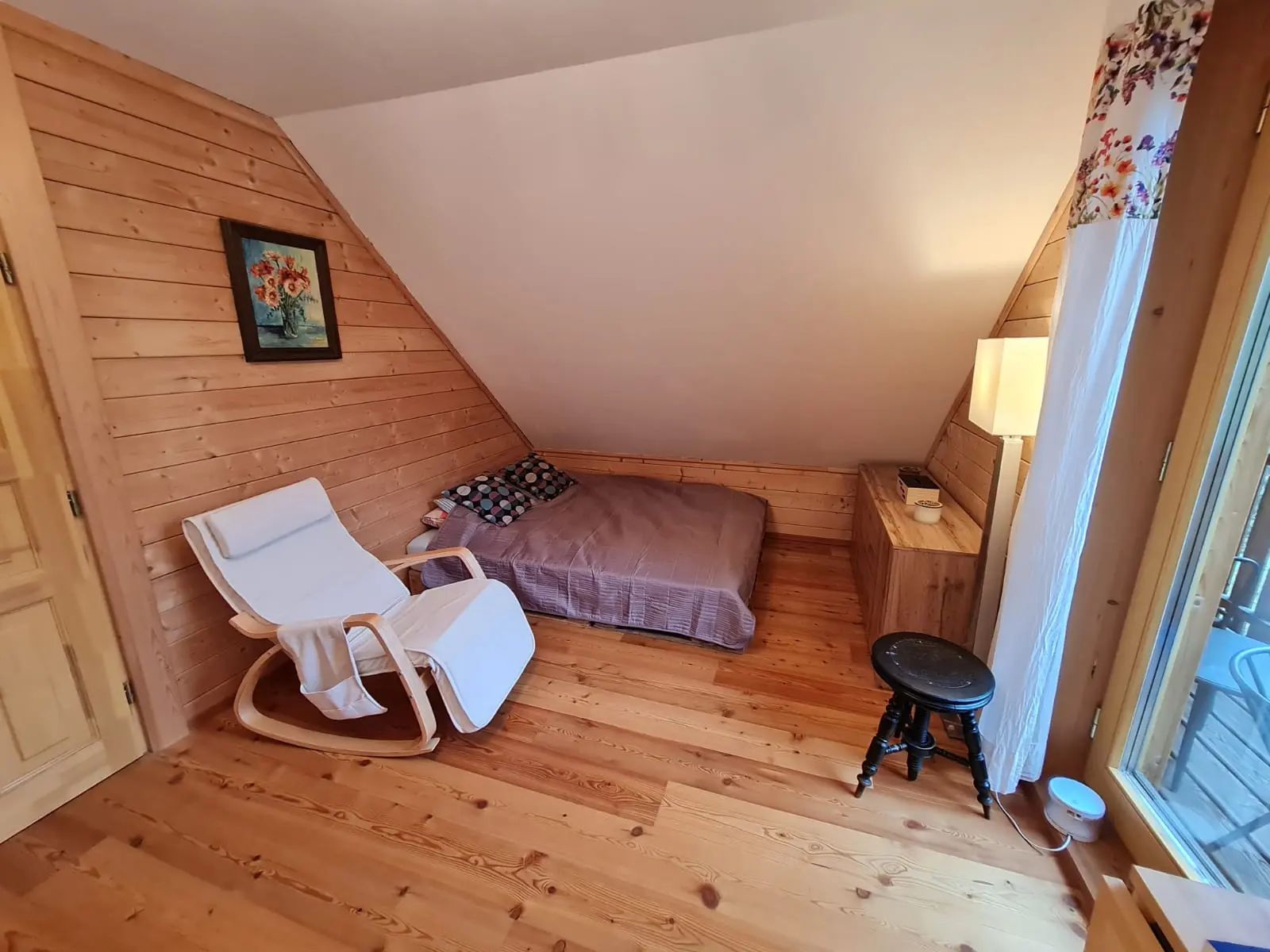 Útulný pokoj s dřevěnými stěnami, postelí, bílým houpacím křeslem a malým stolkem.