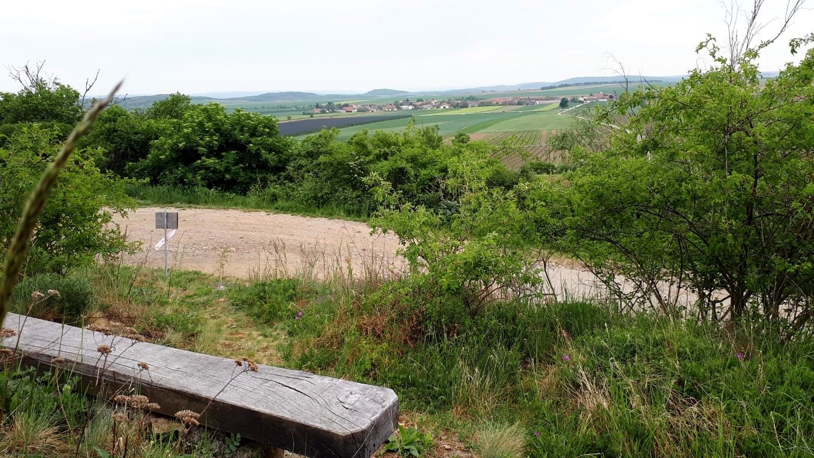 Krajina s lavičkou, stromy a poli v dálce.
