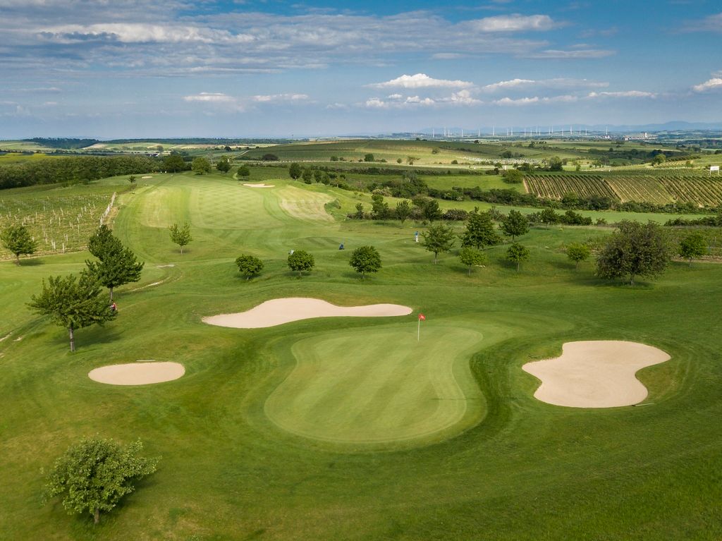 Letecký pohled na golfové hřiště se zelenými plochami, písečnými bunkry a okolními poli.
