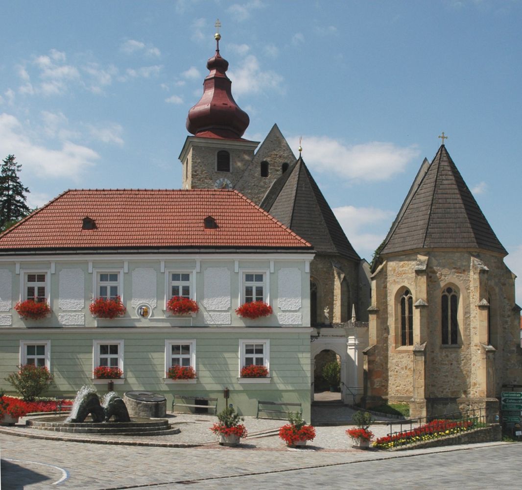 Historická budova s červenou střechou a kostelem v pozadí na náměstí Maria-Anzbach.