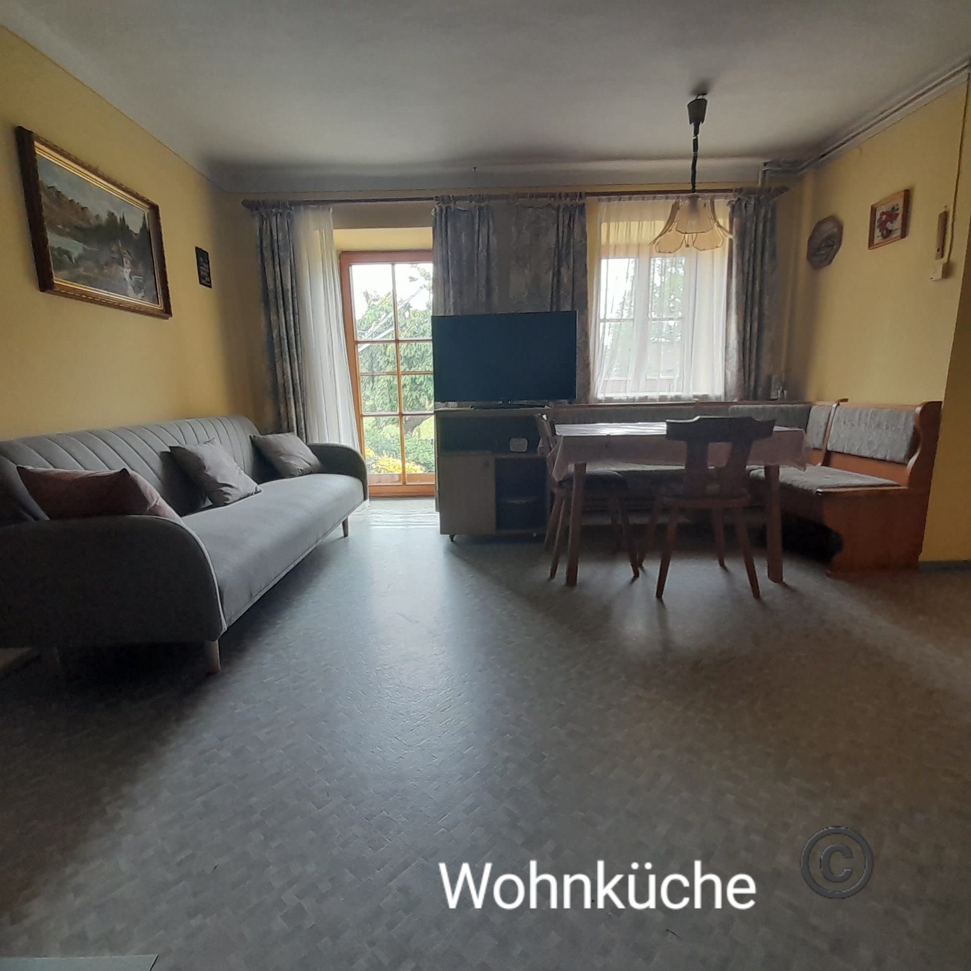 Obývací pokoj s kuchyní, pohovkou, jídelním stolem a TV před velkými okny.