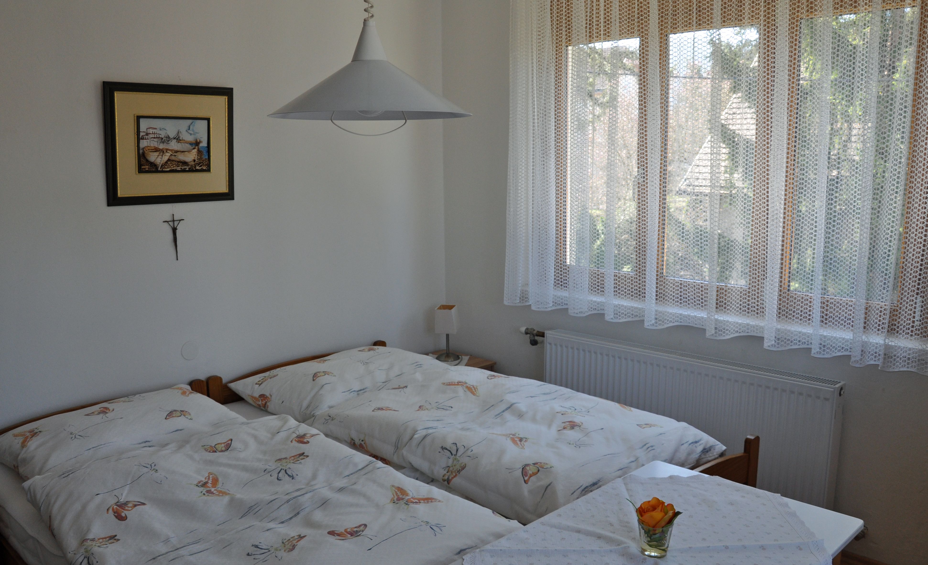 Útulná ložnice se dvěma samostatnými lůžky, obrazem na stěně a oknem s krajkovým závěsem.