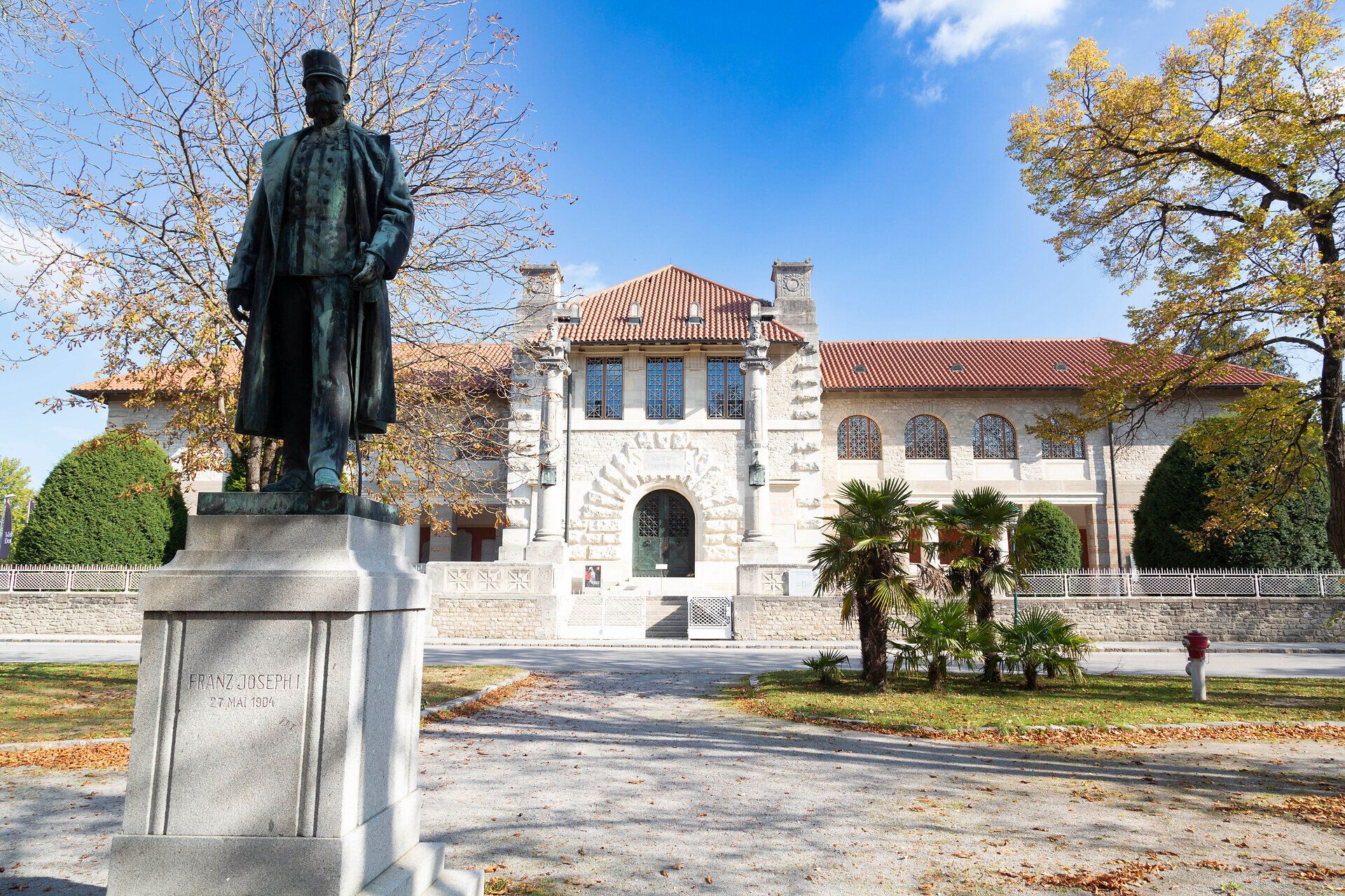 Majestátní socha stojí hrdě před impozantní budovou, obklopená barevným podzimním listím. Jemné sluneční paprsky zalévají scenérii teplým světlem a vybízejí k prozkoumání krásy okolí.