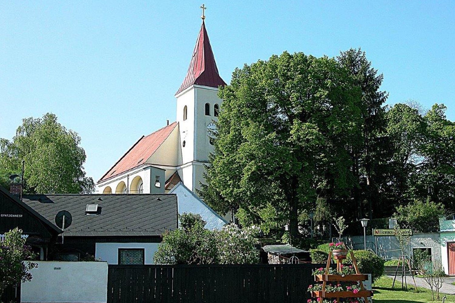 Kostel s červenou střechou a věží, obklopený stromy a domy.