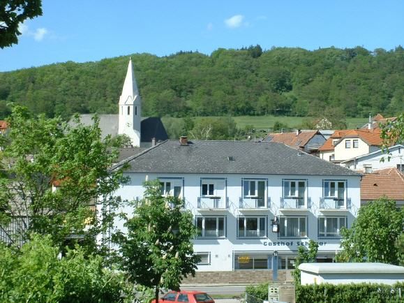 Dvoupatrová budova s nápisem "Gasthof Schauer" před zalesněným kopcem a kostelem se špičatou věží.