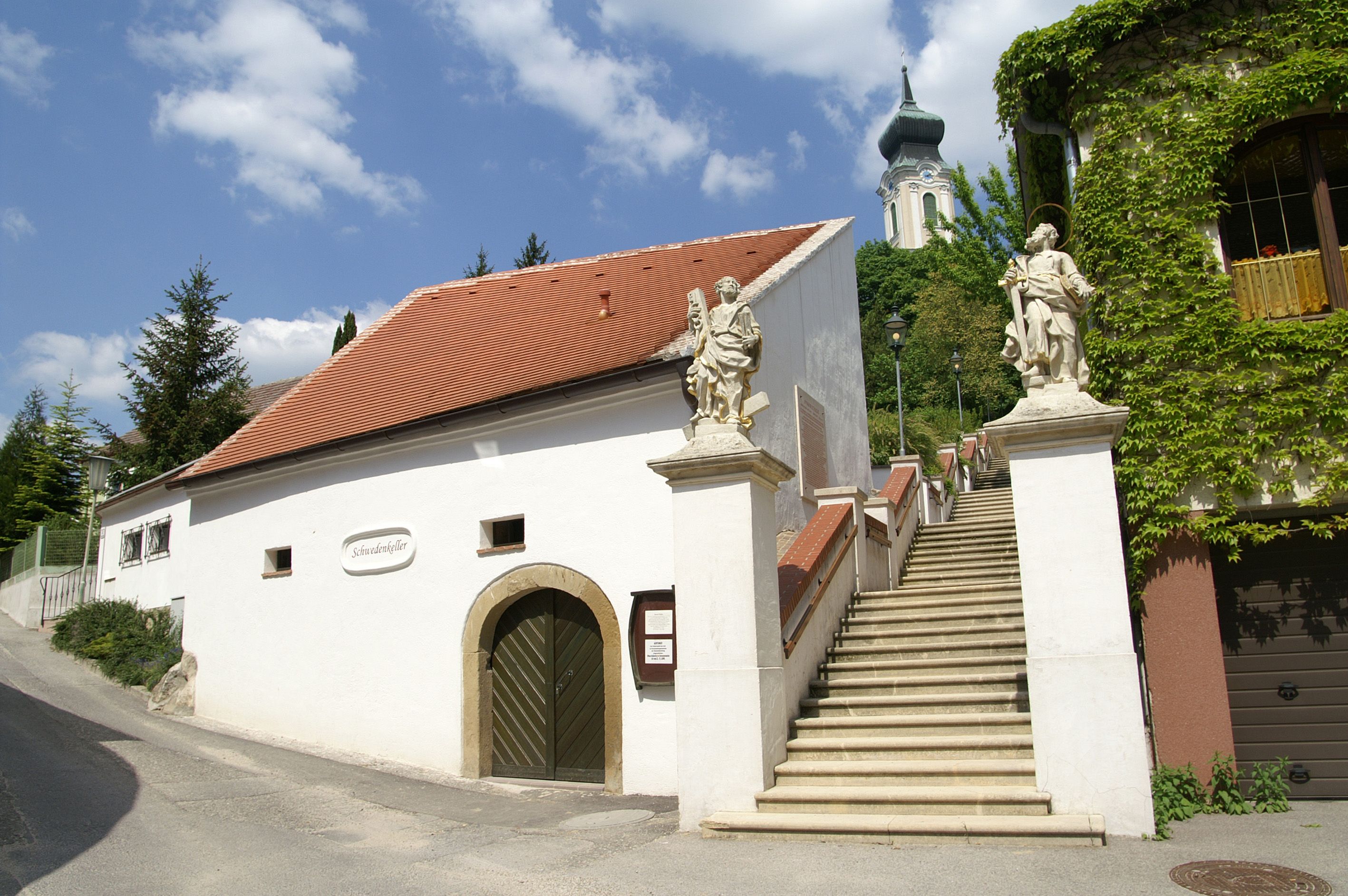 Bílá budova s červenou střechou a schodištěm se sochami, v pozadí kostel.