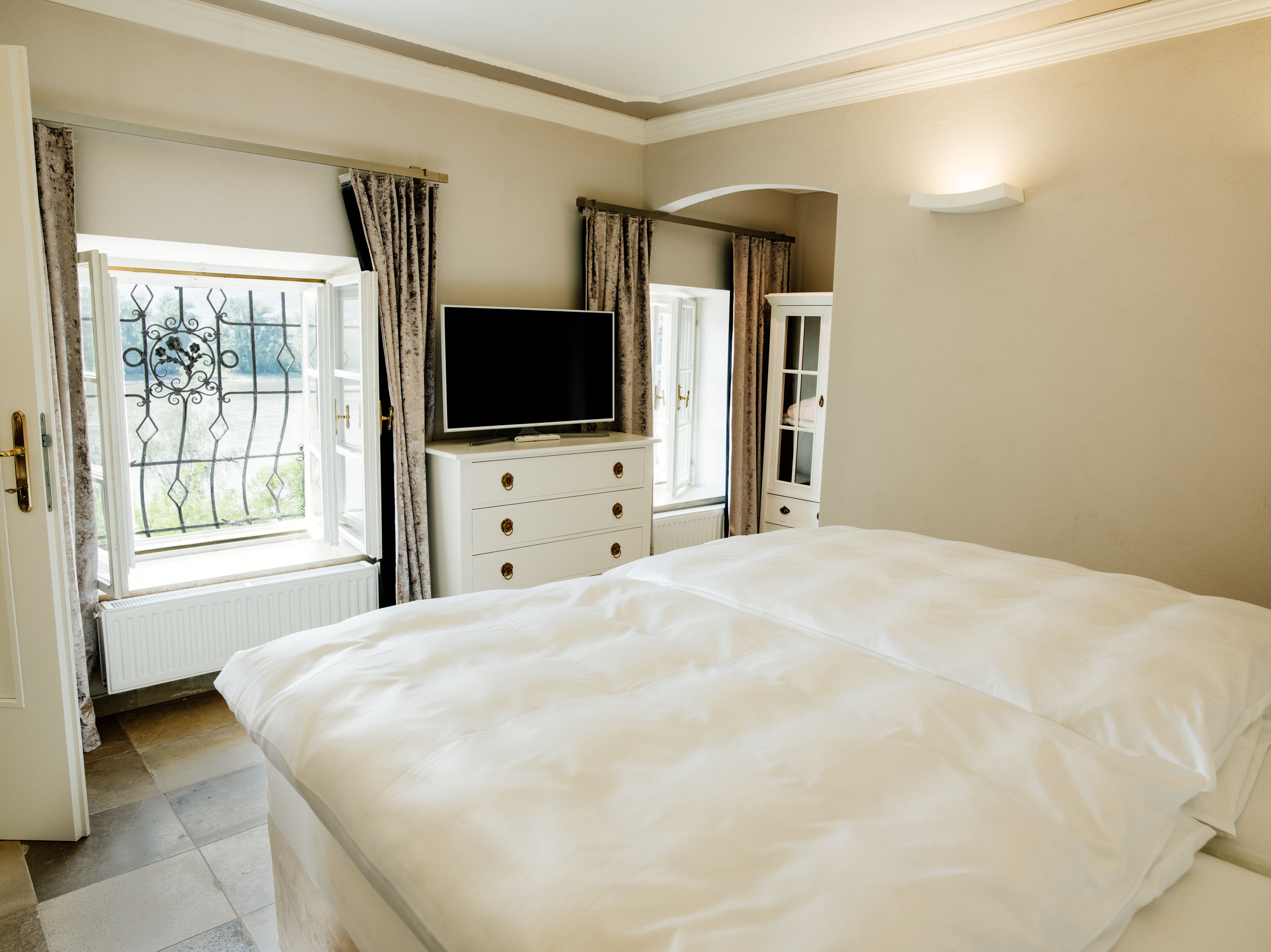 Světlá ložnice s manželskou postelí, TV, komodou a dvěma okny se závěsy.