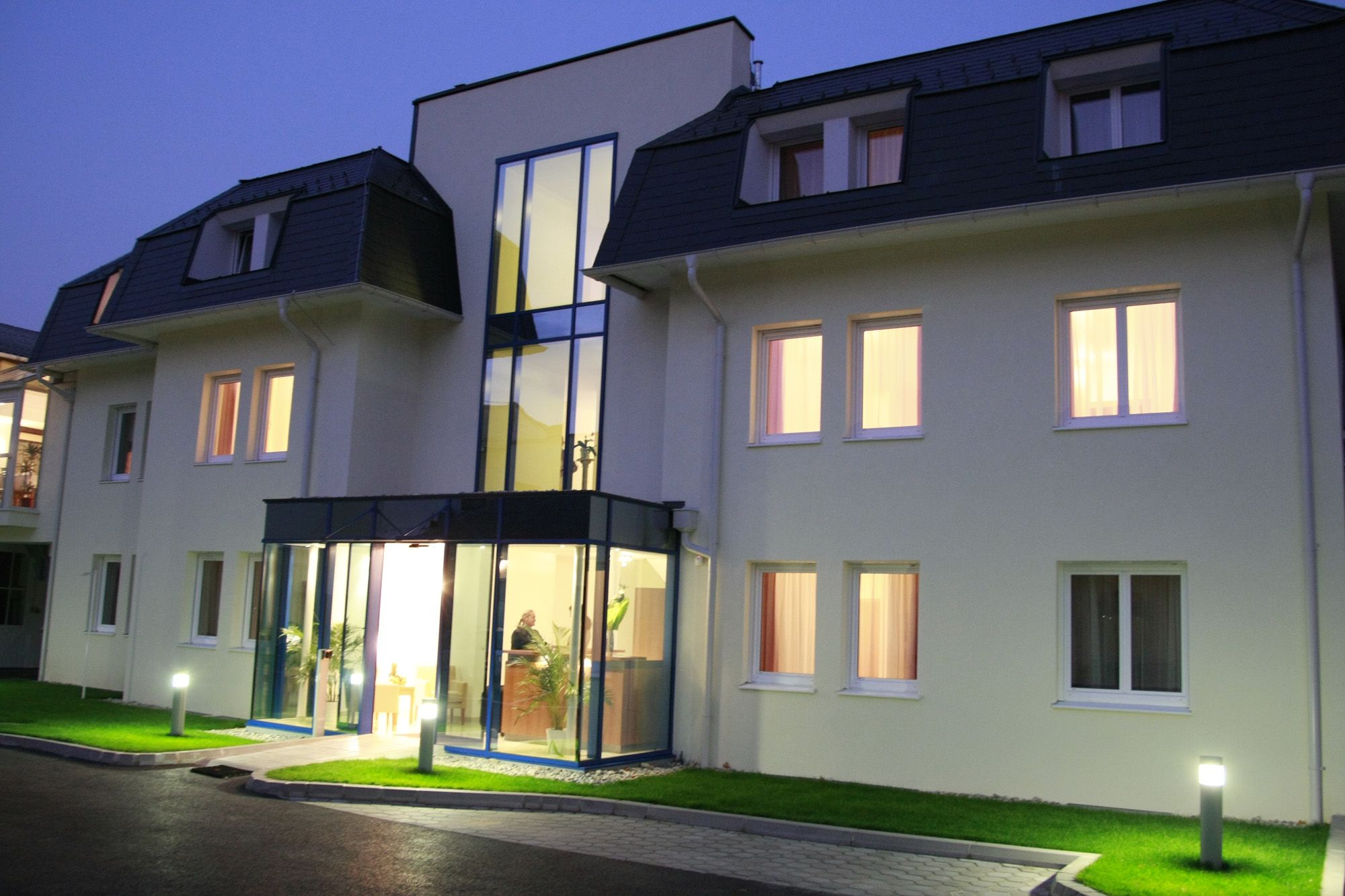Vstupní prostor moderního hotelu za soumraku s osvětlenými okny a skleněnou fasádou.