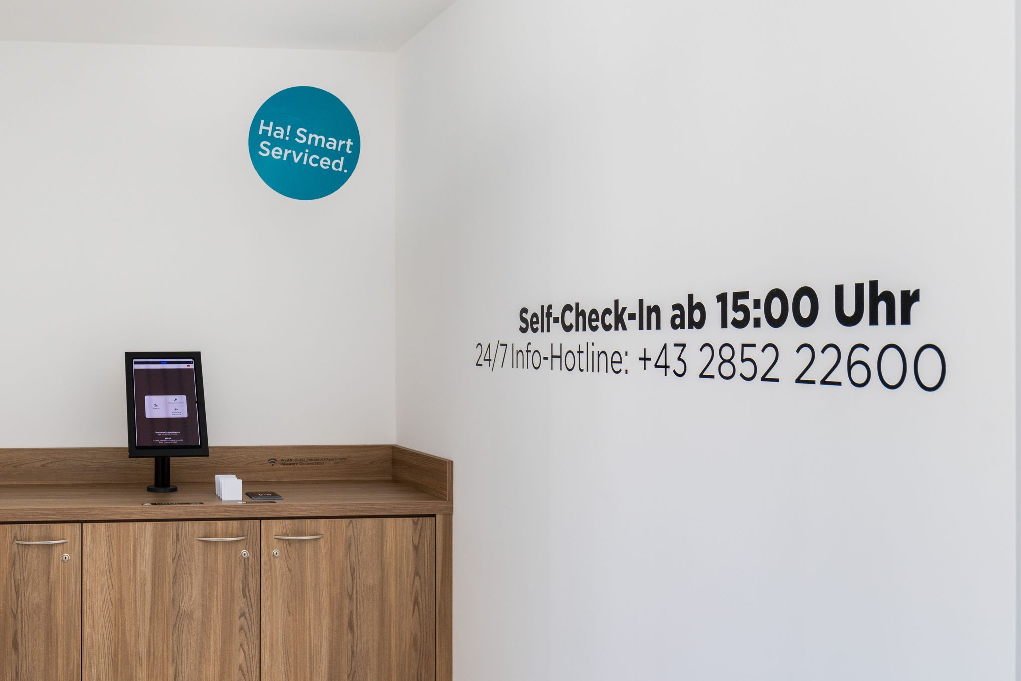 Prostor pro samoobslužnou registraci s obrazovkou a telefonním číslem na nepřetržitou linku pomoci vyvěšeným na zdi.