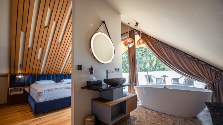 Moderní hotelový pokoj s dřevěným stropem, volně stojící vanou a balkonem.