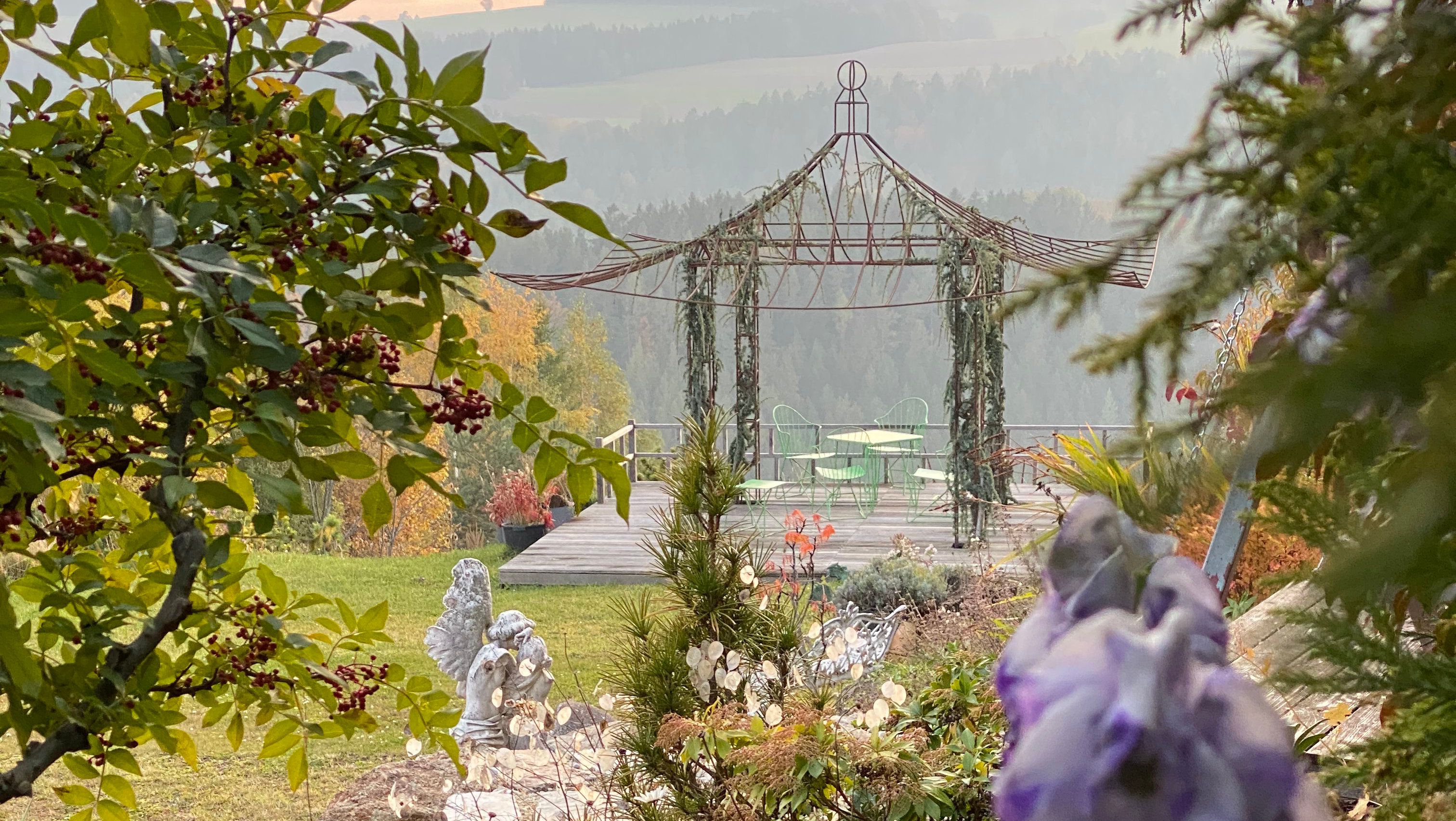 Romantická zahrada s pavilonem, zelenými židlemi a stolem, obklopená stromy a květinami.