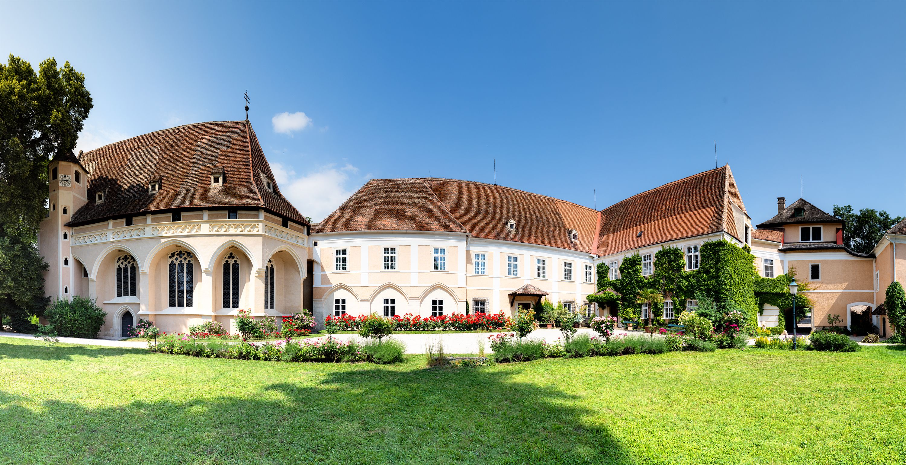 Panoramatický pohled na zámek Schrattenthal s upravenou zahradou a kvetoucími růžemi v popředí.