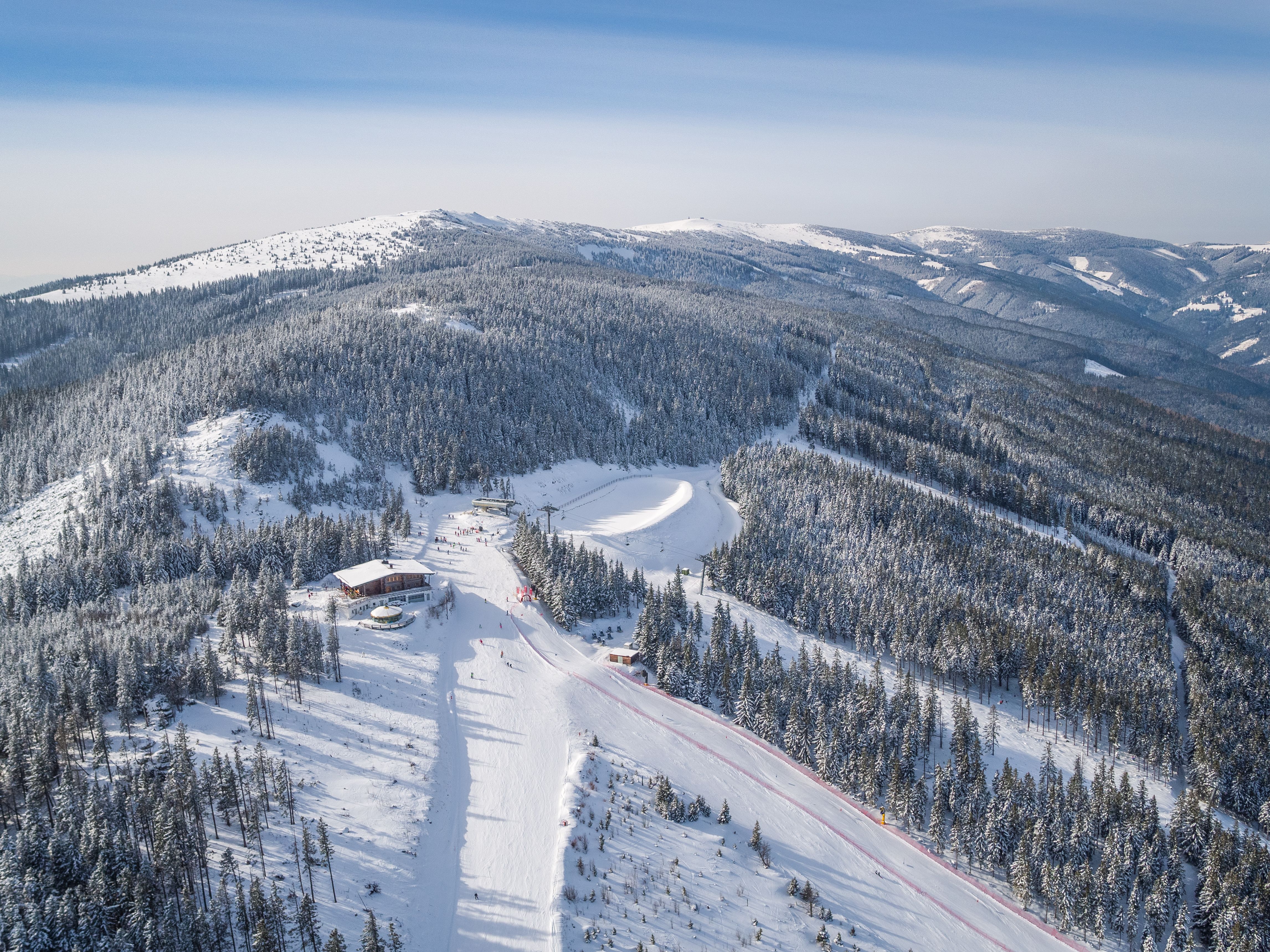 Lyžařský areál se sedačkovou lanovkou a lyžaři, obklopený zasněženými stromy a kopci.