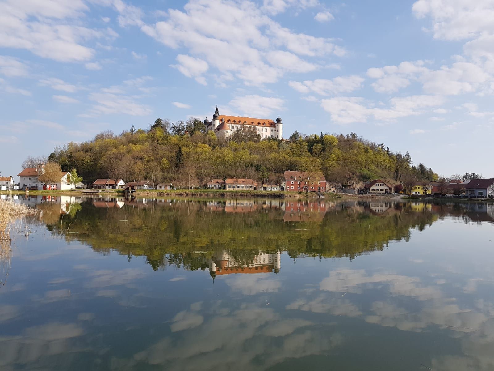 Hrad na kopci se odráží v klidném jezeře obklopeném stromy a malými domky.