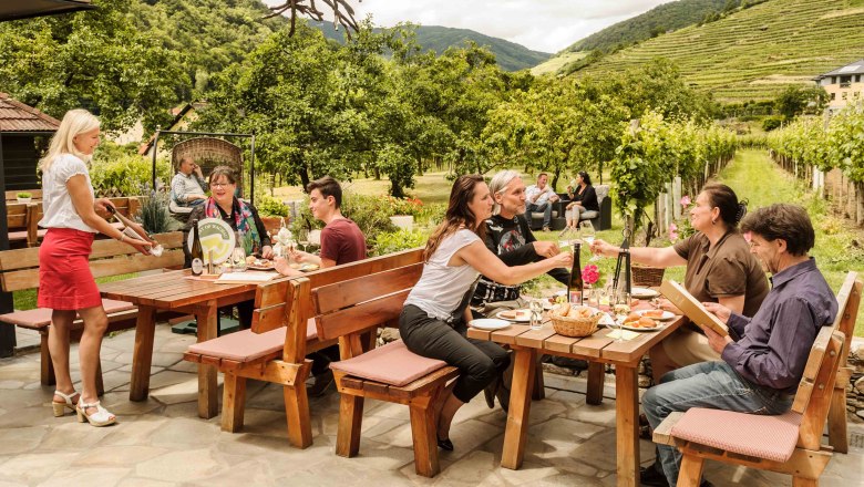 Lidé sedí u dřevěných stolů venku, obklopeni vinicemi a stromy, a servírka jim podává nápoje.