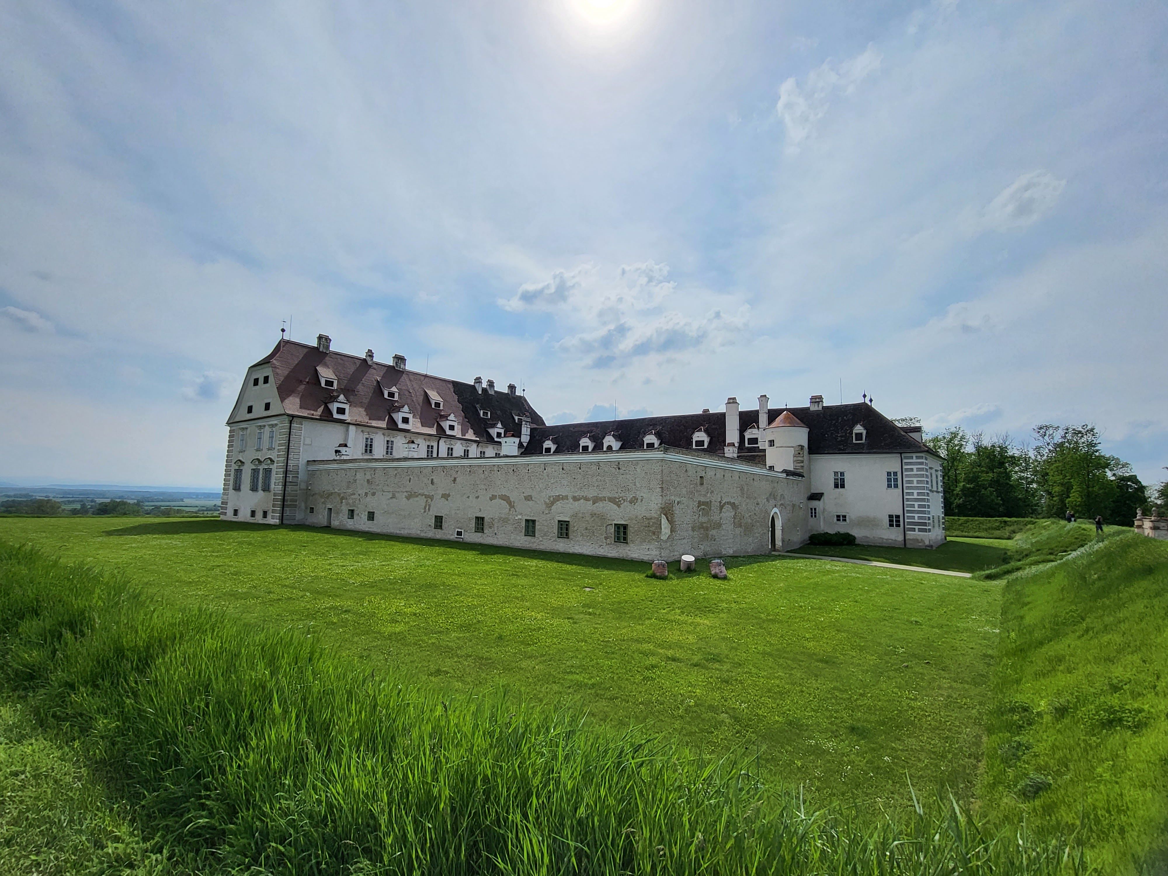 Na zelené louce pod zataženou oblohou stojí velký historický hrad s bílými zdmi a červenými střechami.