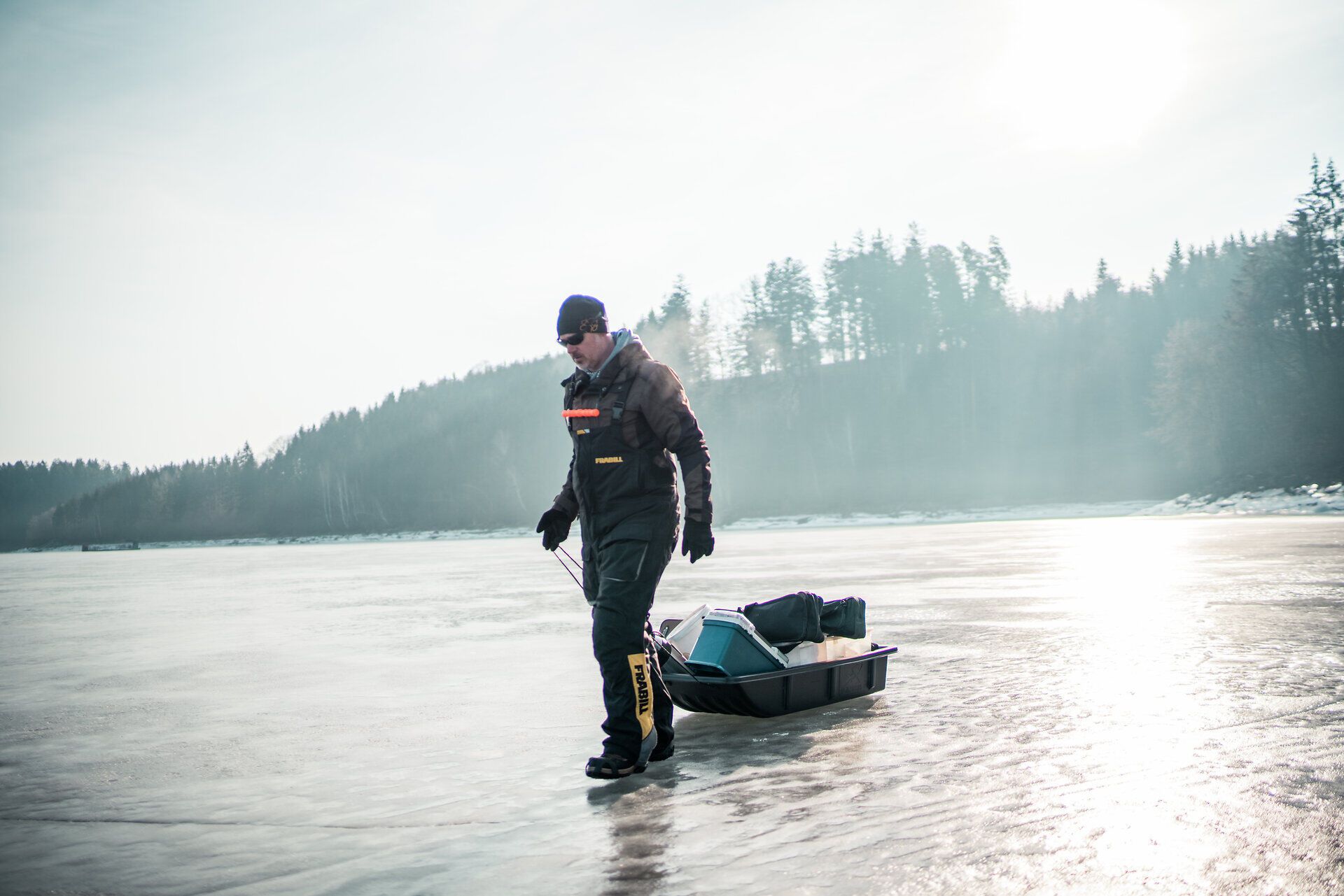 Ein Fischer zieht mit ruhigen Schritten seinen Schlitten über die glitzernde Eisfläche des Stausees. Die frostige Luft ist klar und frisch, während die umgebenden Bäume in sanften Nebelschwaden verschwinden. Hier, in der stillen Winterlandschaft, wird die Geduld und die Freude am Eisfischen spürbar.