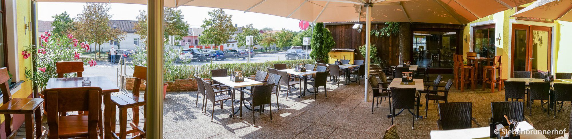 Slunná zahrada pro hosty s prázdnými stoly a židlemi, obklopená rostlinami a velkým slunečníkem.