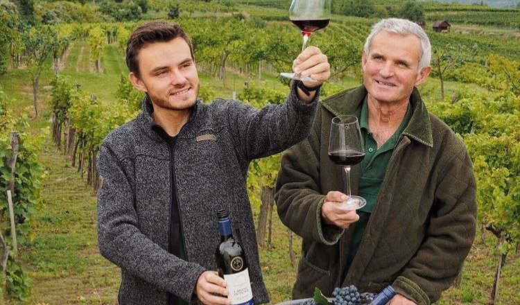 Dva muži na vinici drž&iacute; sklenice s červen&yacute;m v&iacute;nem a usm&iacute;vaj&iacute; se. Jeden z nich drž&iacute; l&aacute;hev v&iacute;na, v pozad&iacute; je vidět vinn&aacute; r&eacute;va a kopce.