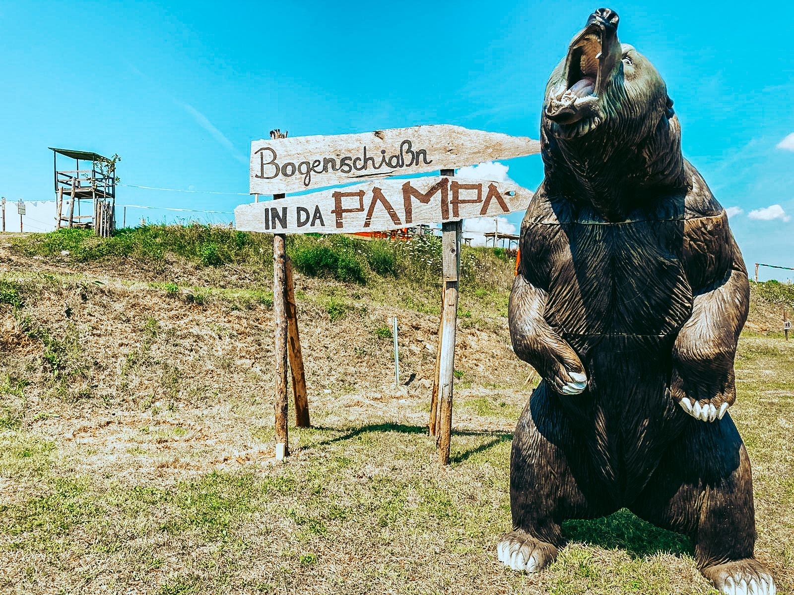 Velký model medvěda vedle cedule s nápisem "Bogenschiaßn in da Pampa" na louce.