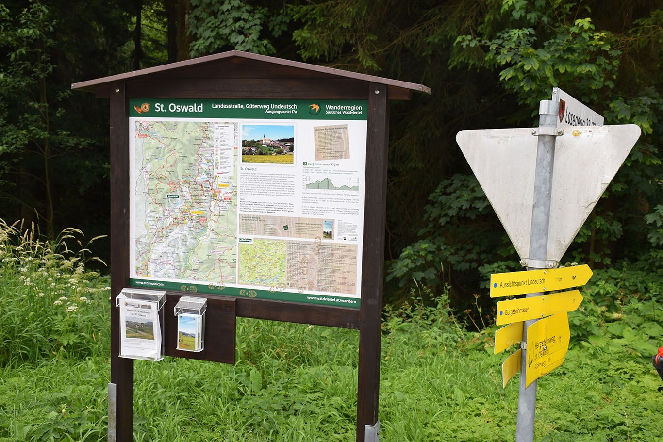 Turistický rozcestník a informační tabule v obci St. Oswald, obklopené lesem a zelení.