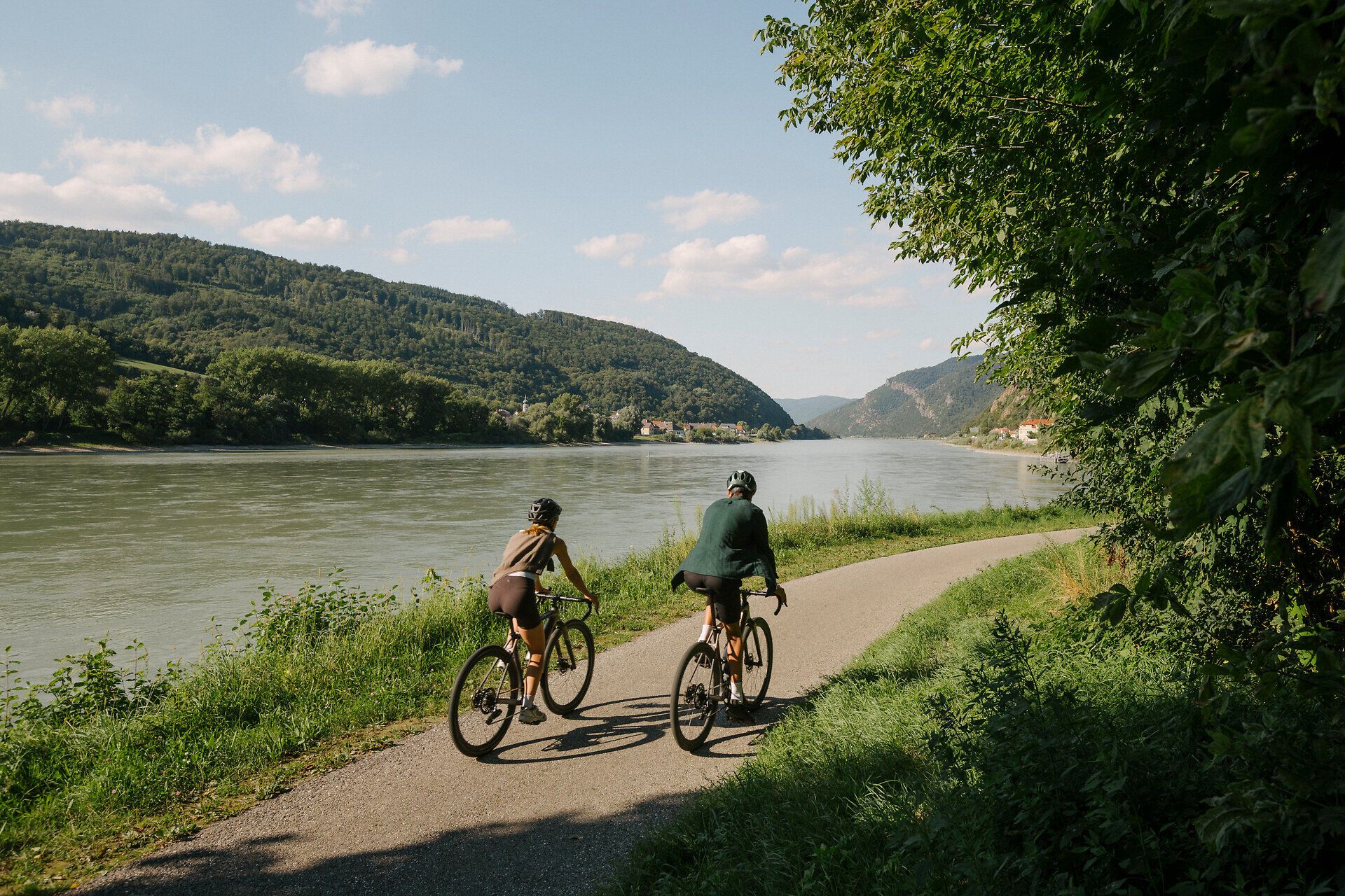 Zwei Radfahrende auf einem asphaltierten Uferweg entlang der Donau; gegenüber liegen bewaldete Hügel, rechts spenden Bäume Schatten.