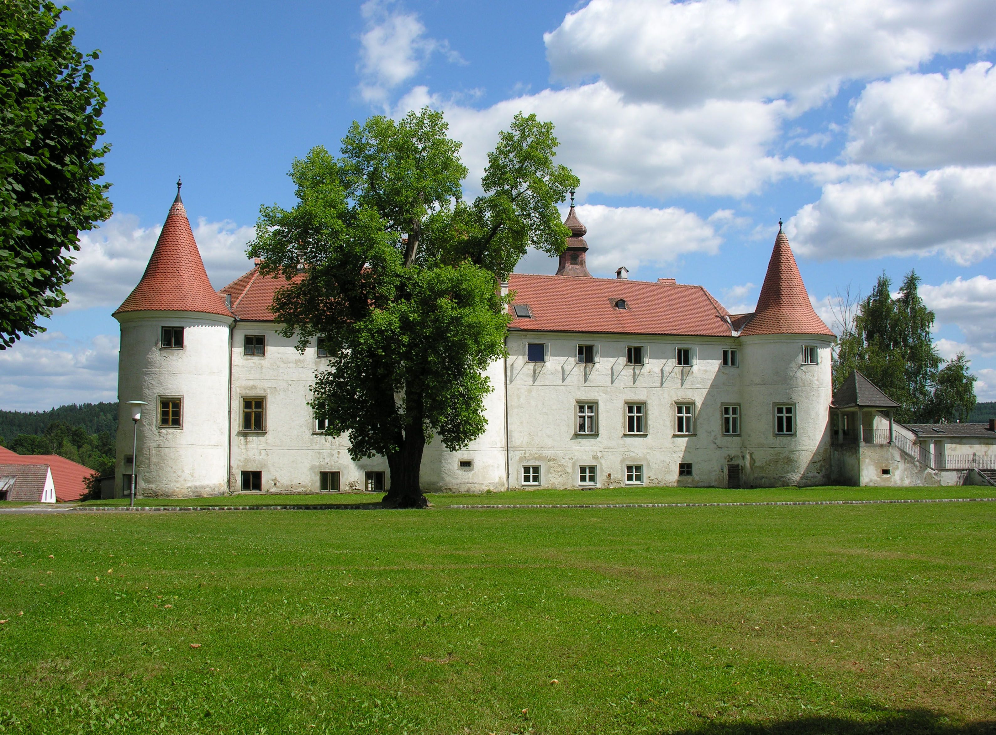 Historický hrad s červenými střechami a věžemi, obklopený zelenými loukami a stromy, pod modrou oblohou s bílými mraky.