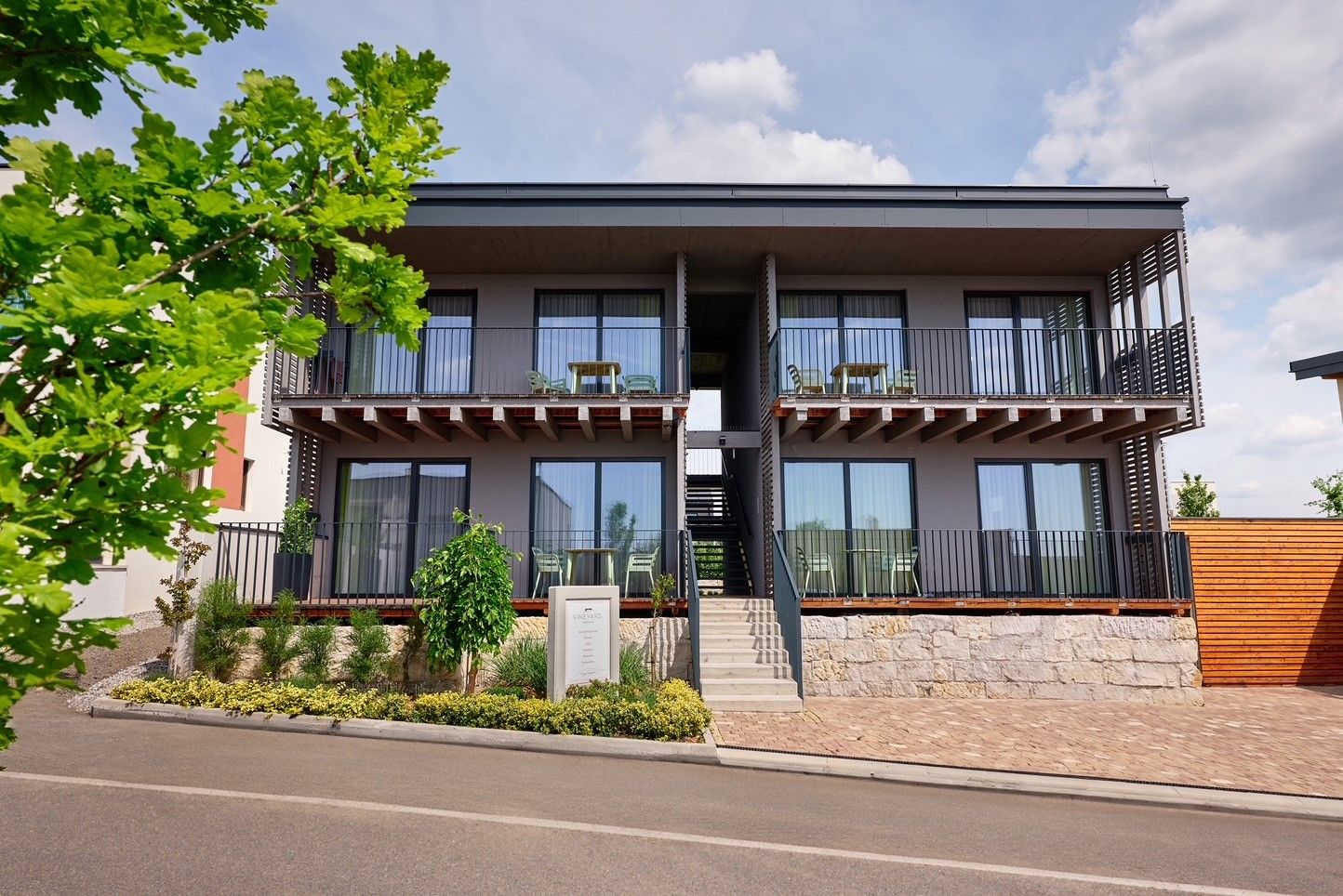 Moderní dvoupodlažní apartmánový komplex s balkony a schodištěm, obklopený rostlinami a stromy.