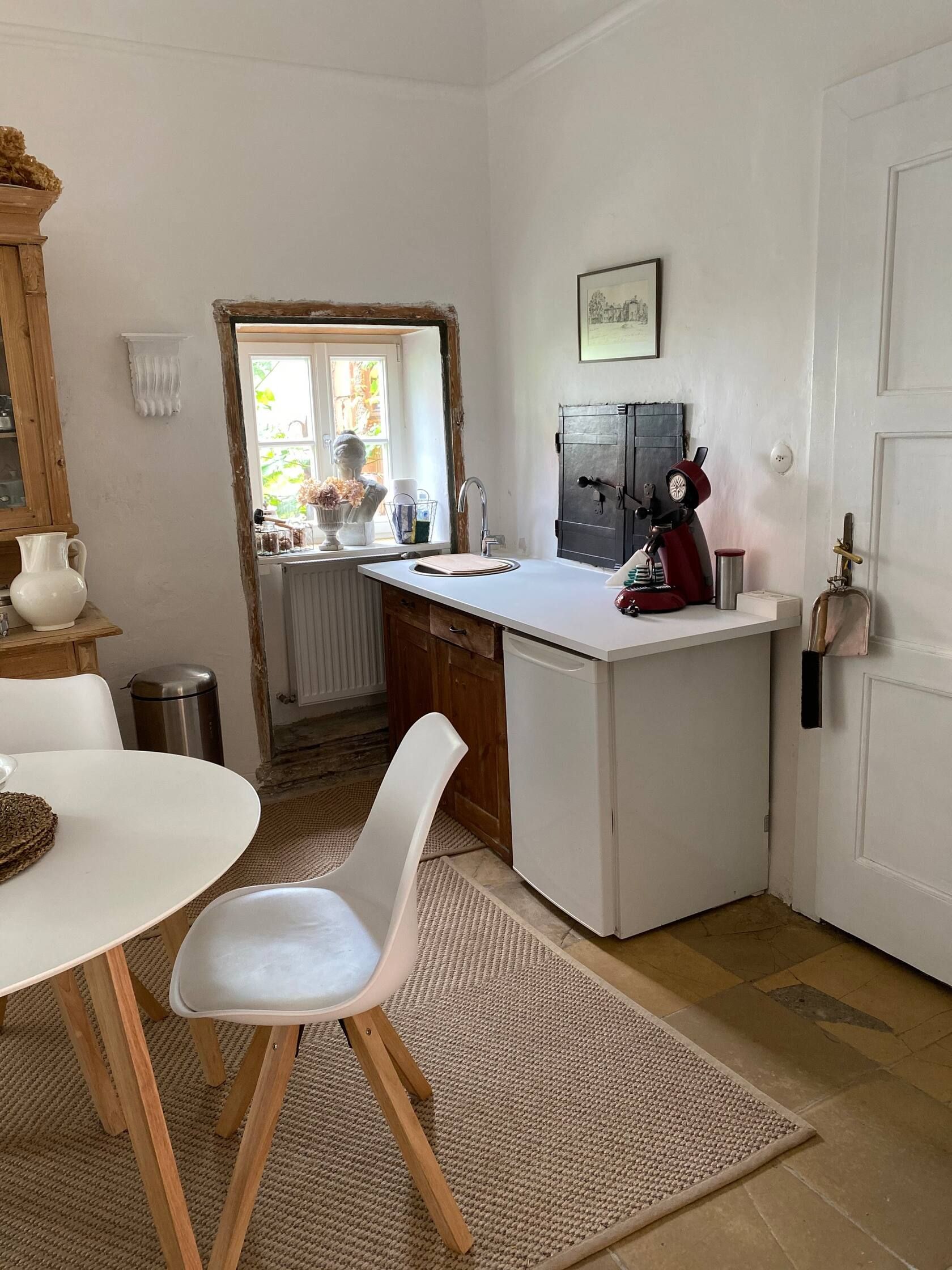 Kuchyňský kout se stolkem, židlí, dřezem a kávovarem. Okna s dekorací a světlé stěny.