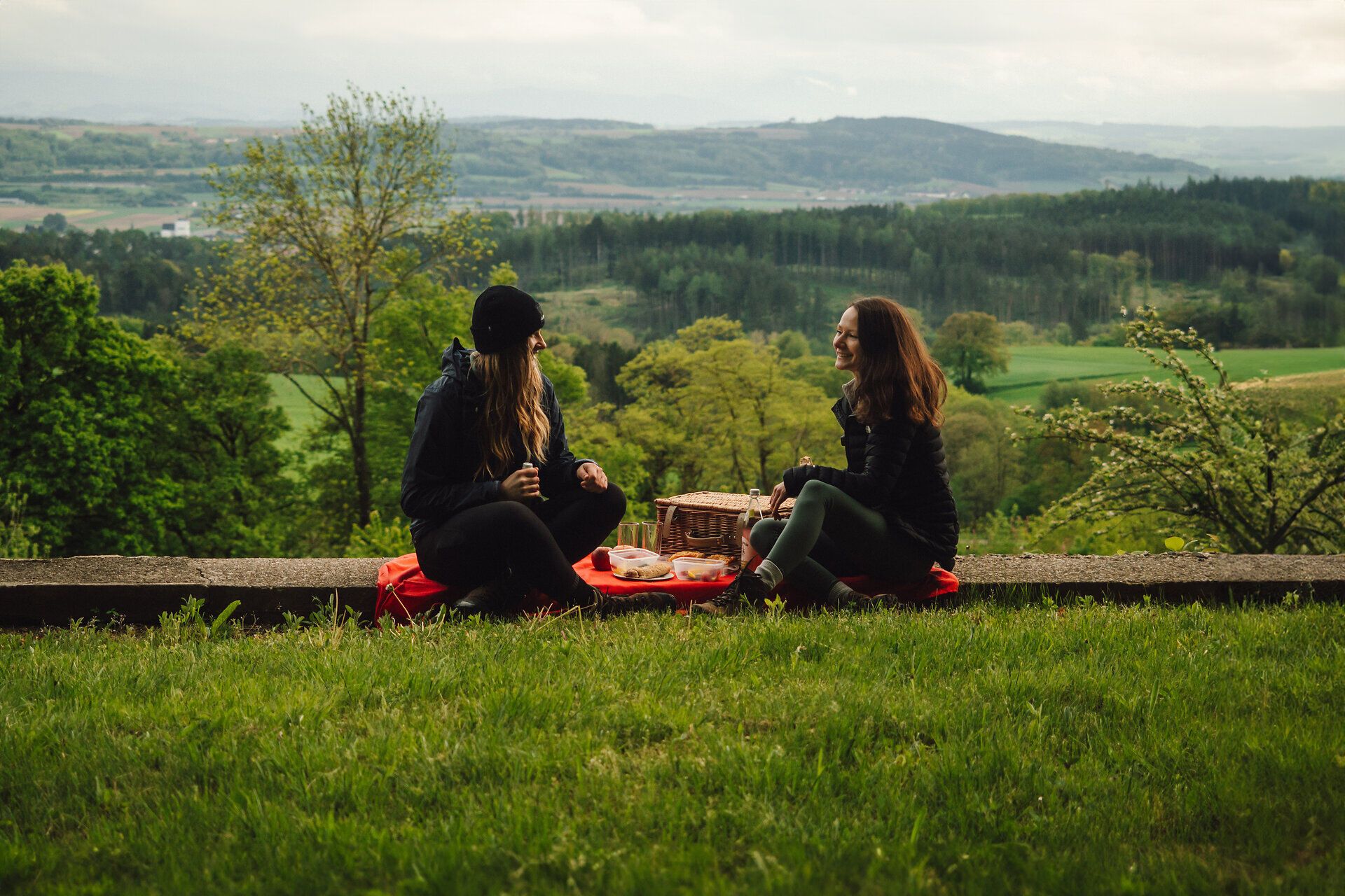 Uprostřed malebné krajiny si dva přátelé užívají pohodový piknik na louce. Scenérie obklopená svěží zelení a kopci vyzařuje poklidnou atmosféru, která vybízí k posezení.