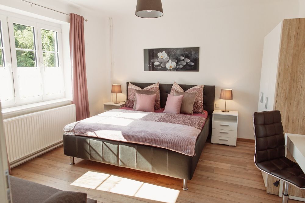 Světlá ložnice s manželskou postelí, dvěma nočními stolky a oknem s růžovými závěsy.