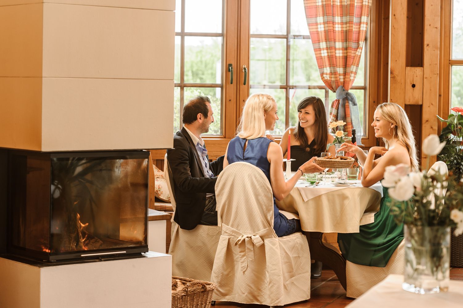 V útulné zimní zahradě s krbem sedí u stolu čtyři osoby.