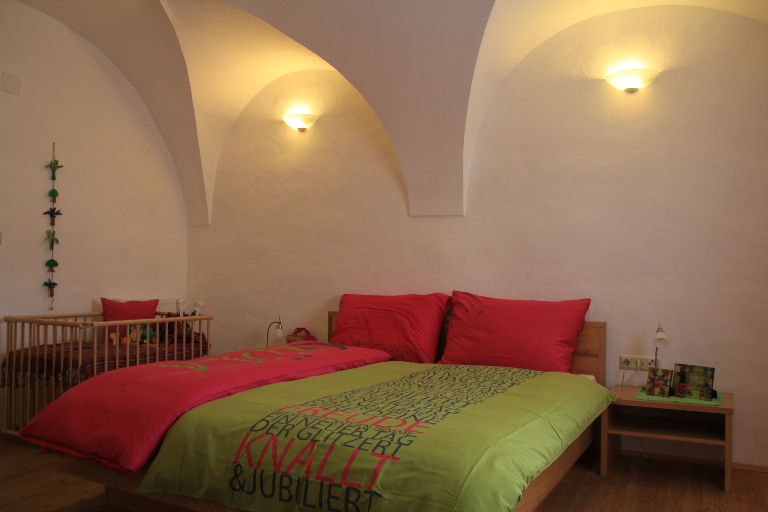 Útulný pokoj pro hosty s manželskou postelí, barevnými peřinami a dětskou postýlkou v pozadí. Stěny jsou bílé a klenuté.