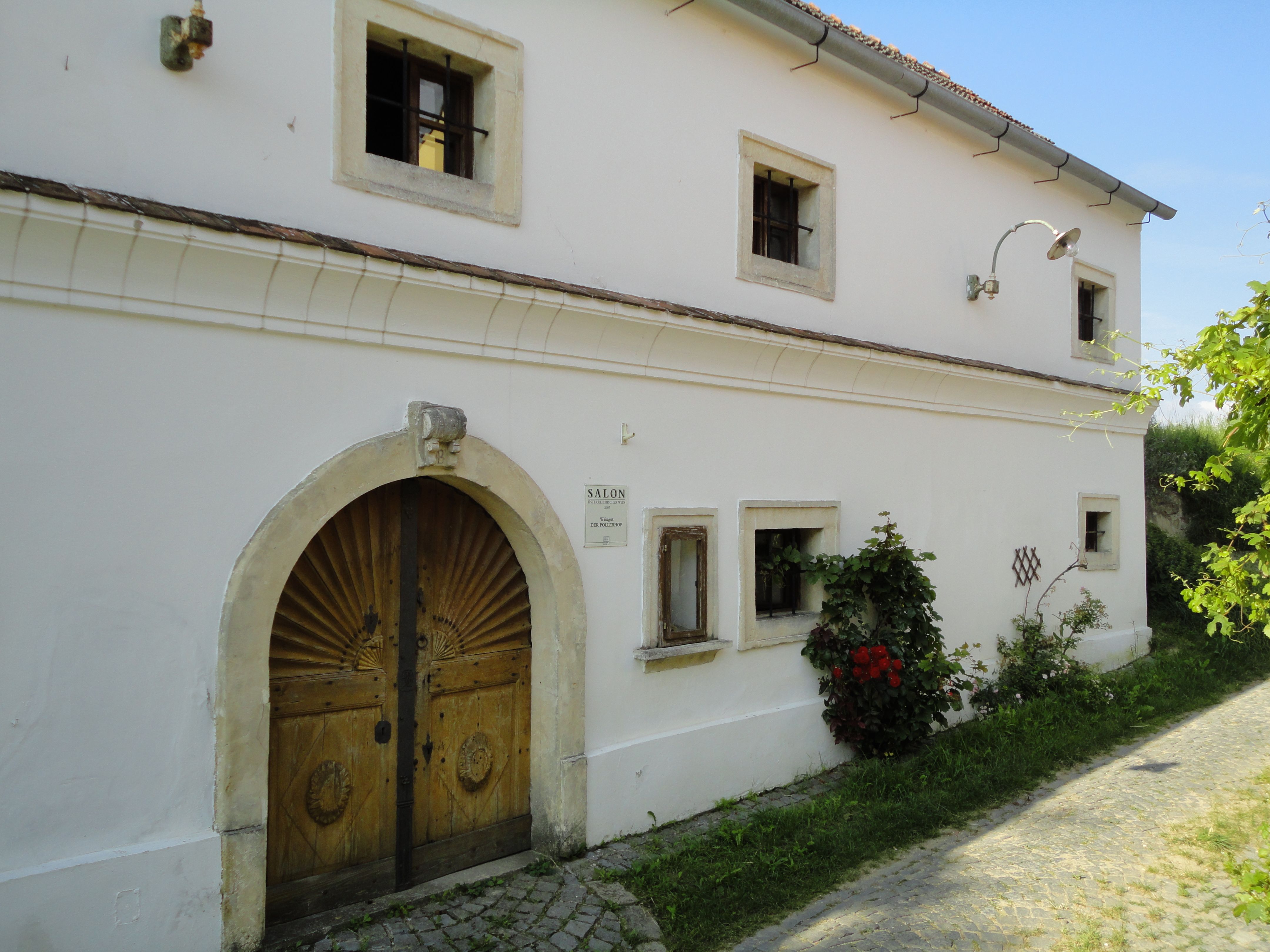Historická budova s dřevěnými dveřmi a malými okny, obklopená rostlinami.