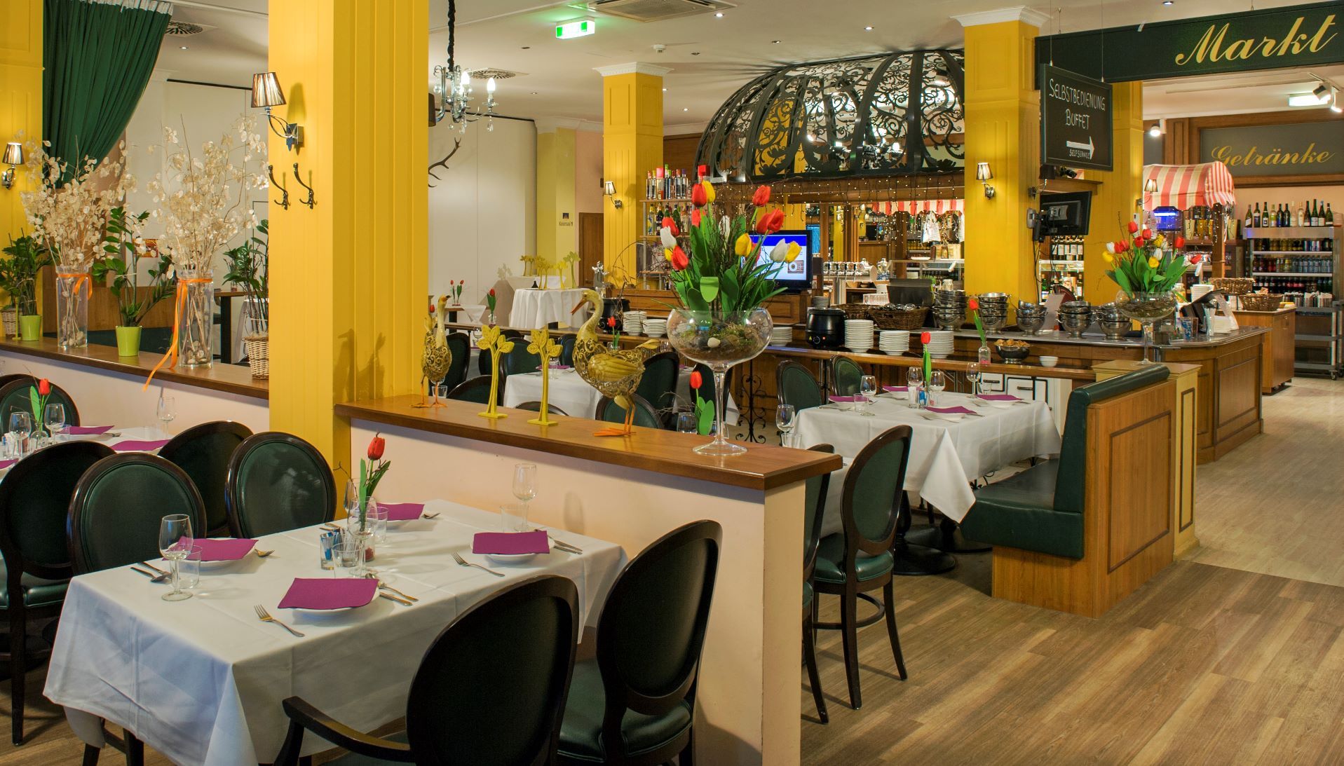 Restaurace se stoly oddělenými polovičními stěnami a žlutými betonovými sloupy. V pozadí je nenápadně vidět kuchyně.