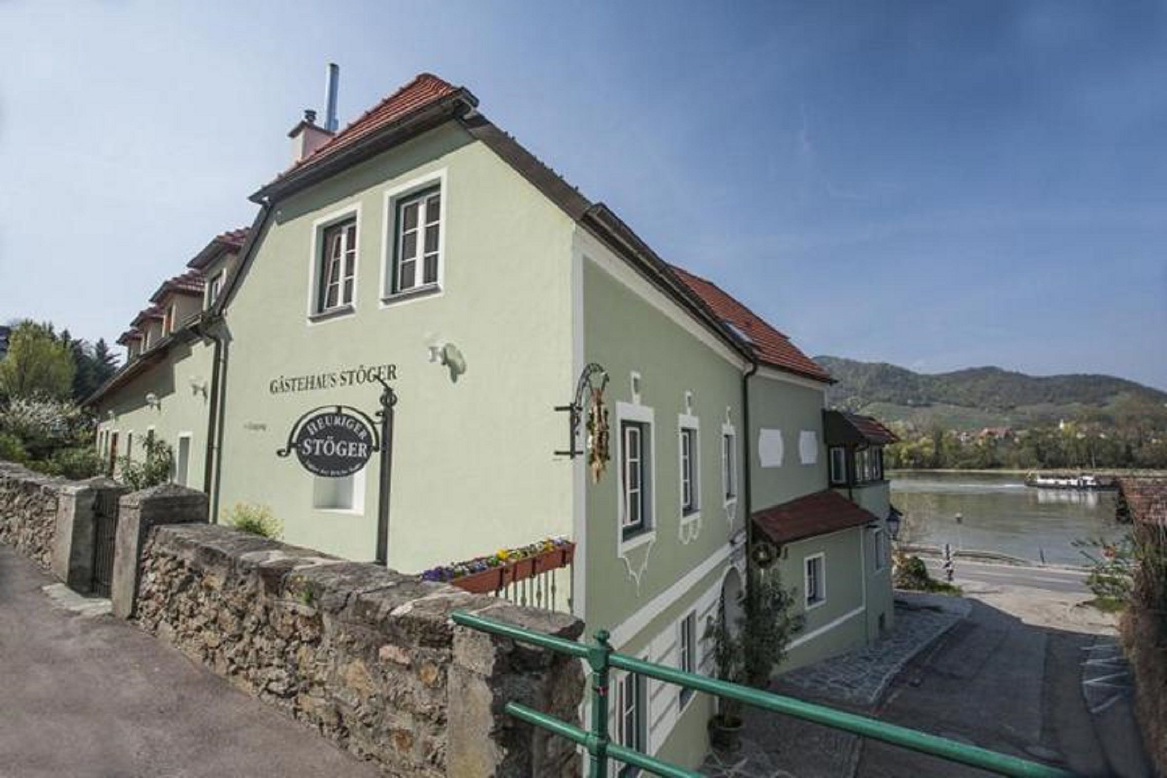 Zelený penzion s označením "Gästehaus Stöger" a "Heuriger Stöger" na břehu řeky.