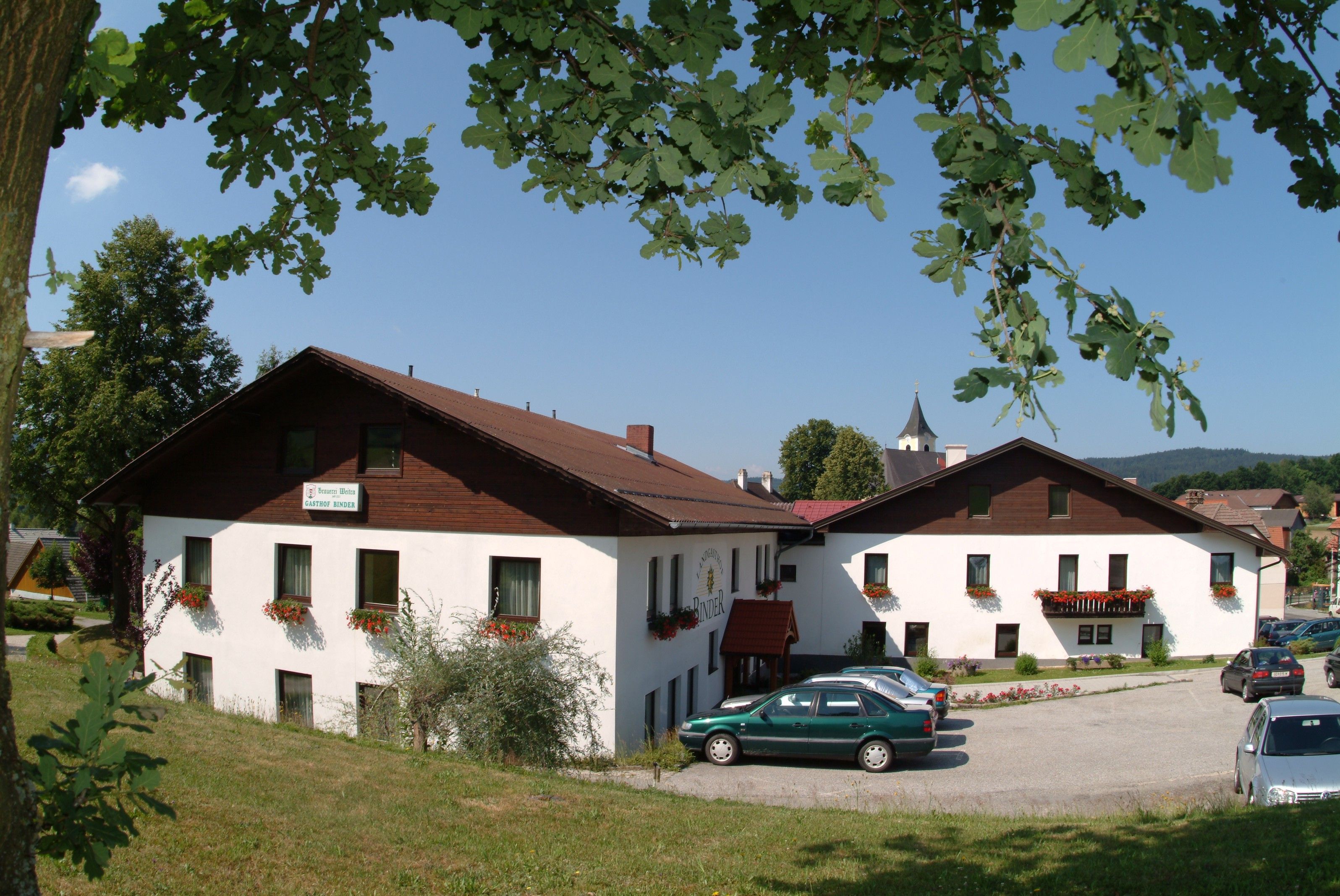 Venkovský hostinec s bílými zdmi a hnědou střechou, obklopený stromy a auty na parkovišti.