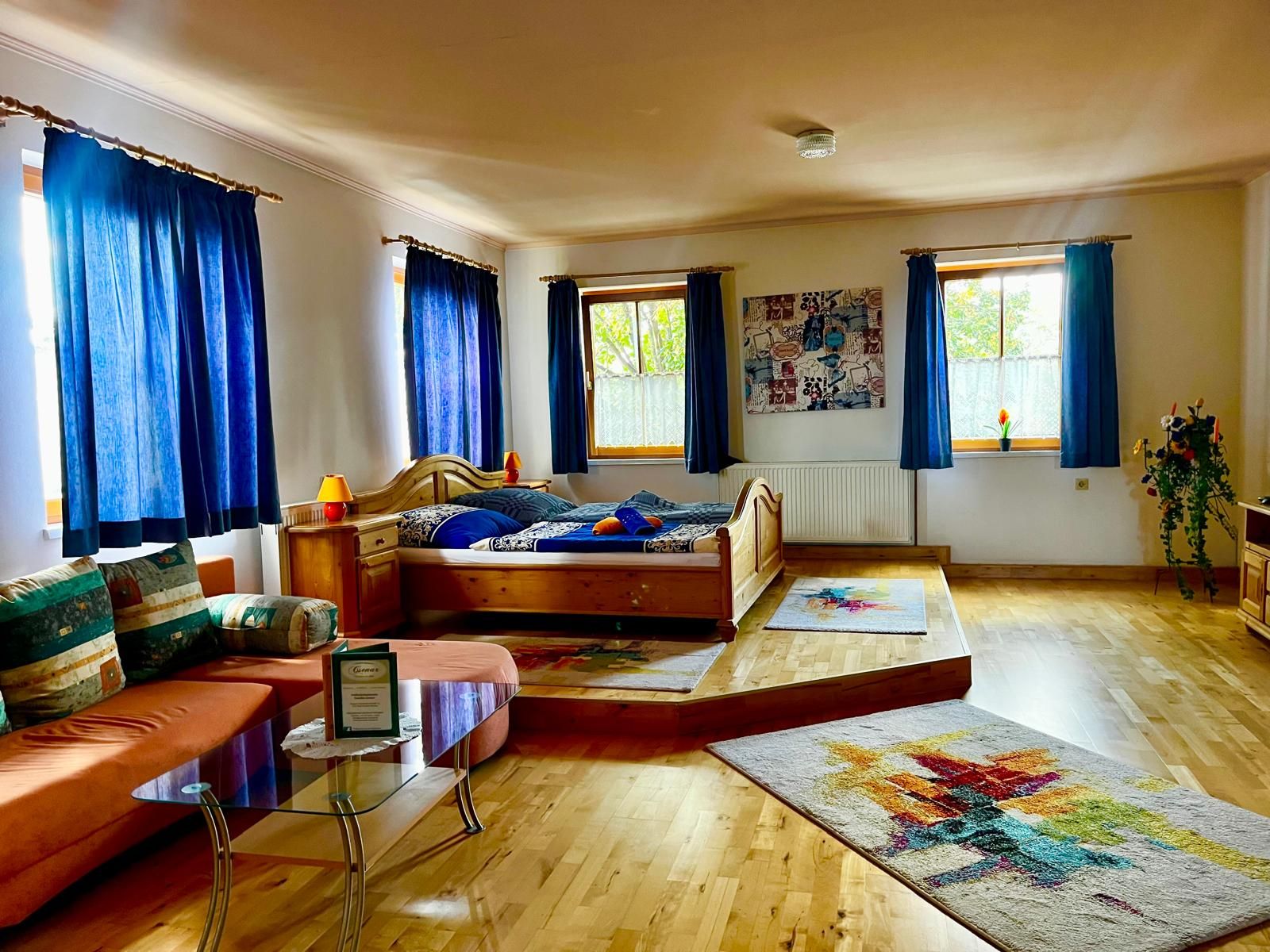 Útulný pokoj s dřevěnou podlahou, manželskou postelí, pohovkou a modrými závěsy.