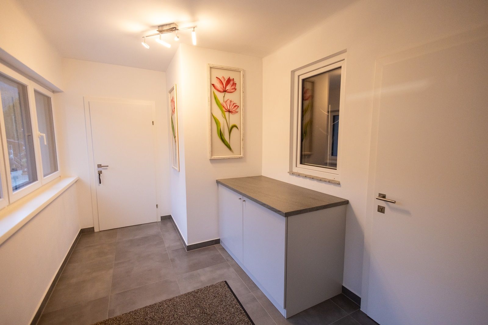 Moderní vstupní hala s bílými stěnami, šedou podlahou, skříní a květinovými obrazy na stěně.