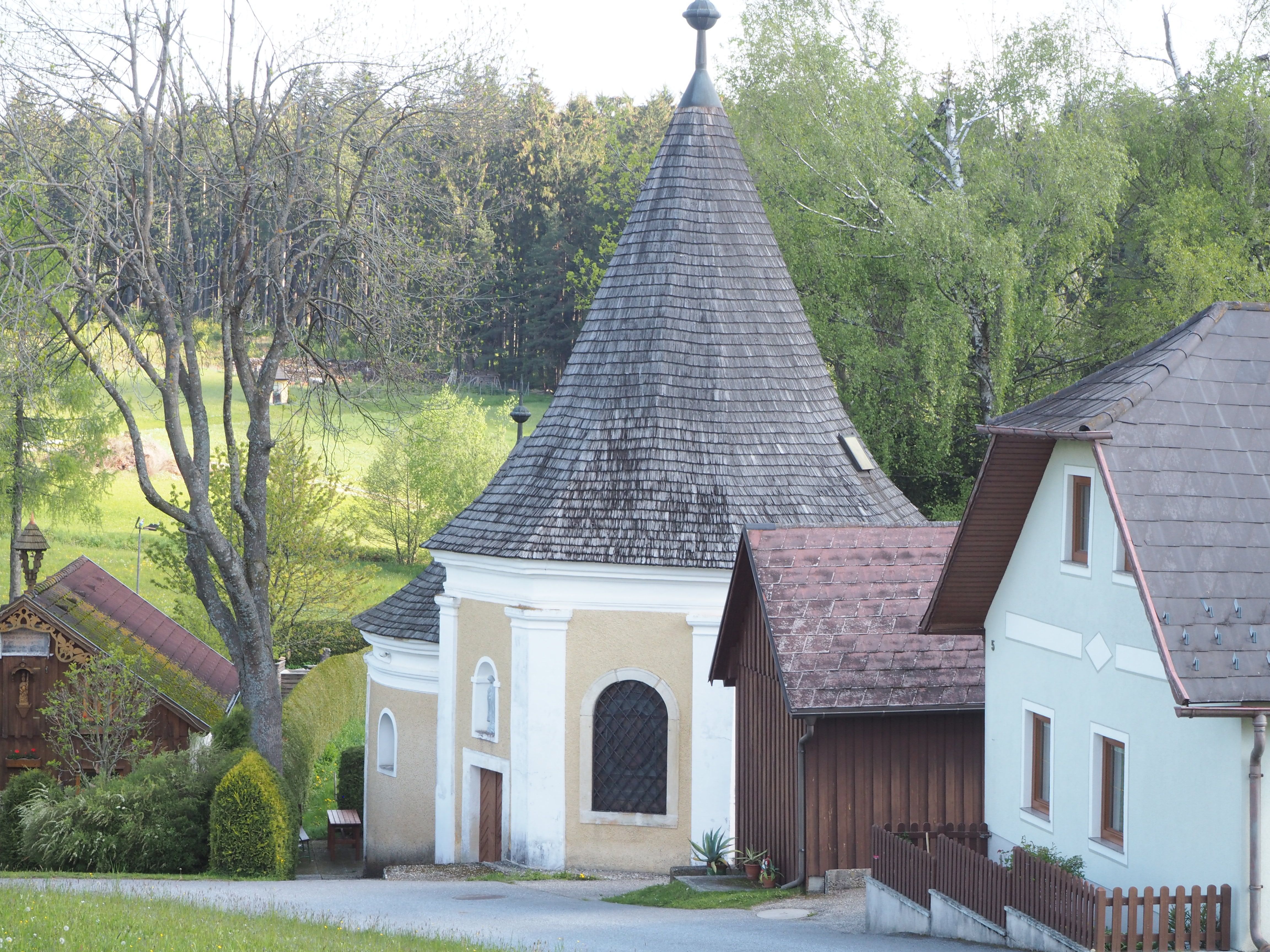 Malá kaple se špičatou střechou, obklopená stromy a domy.